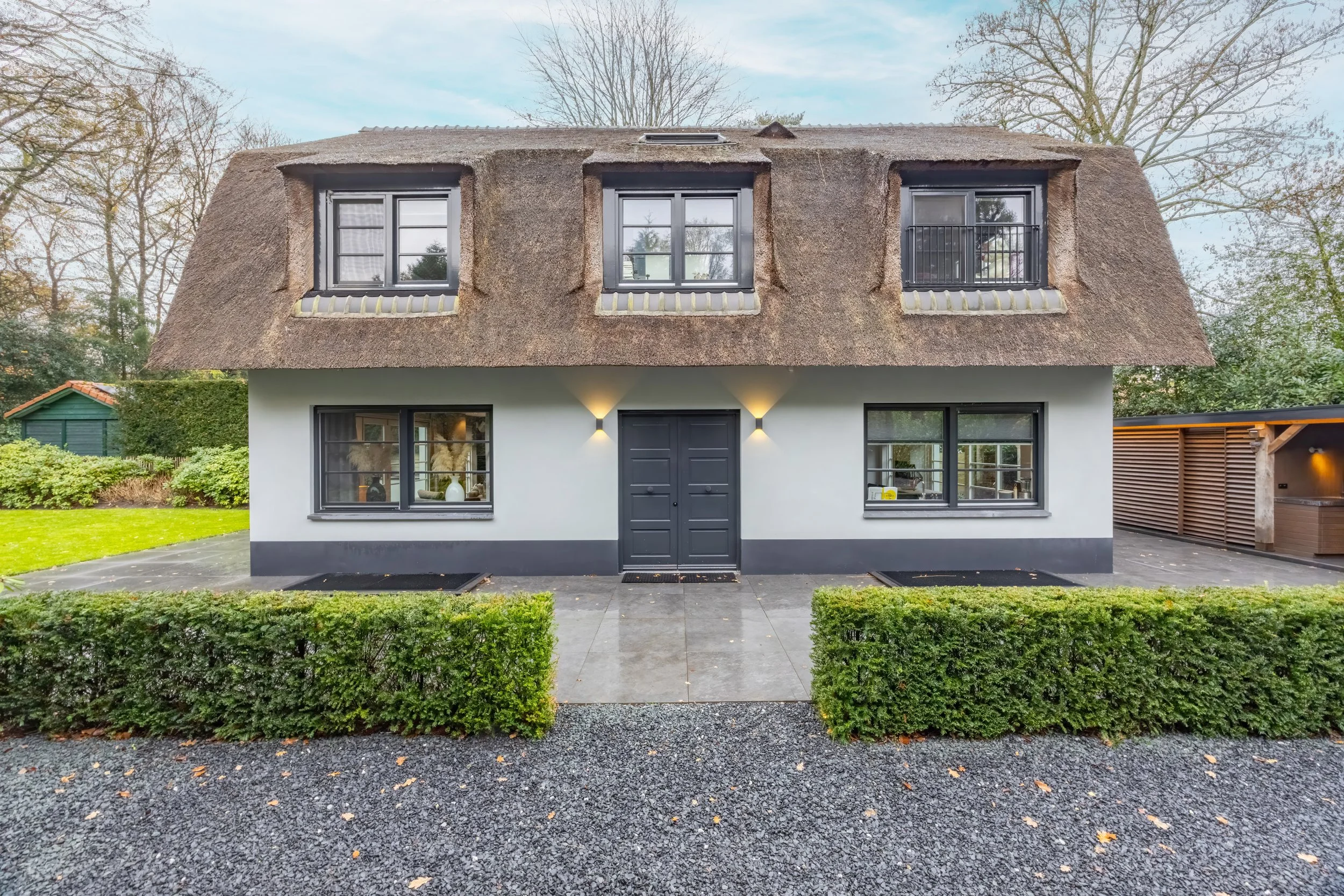Een modern huis met een witte gevel, zwarte ramen en een rieten dak, omgeven door een aanlegsteiger met struiken en een tuin.