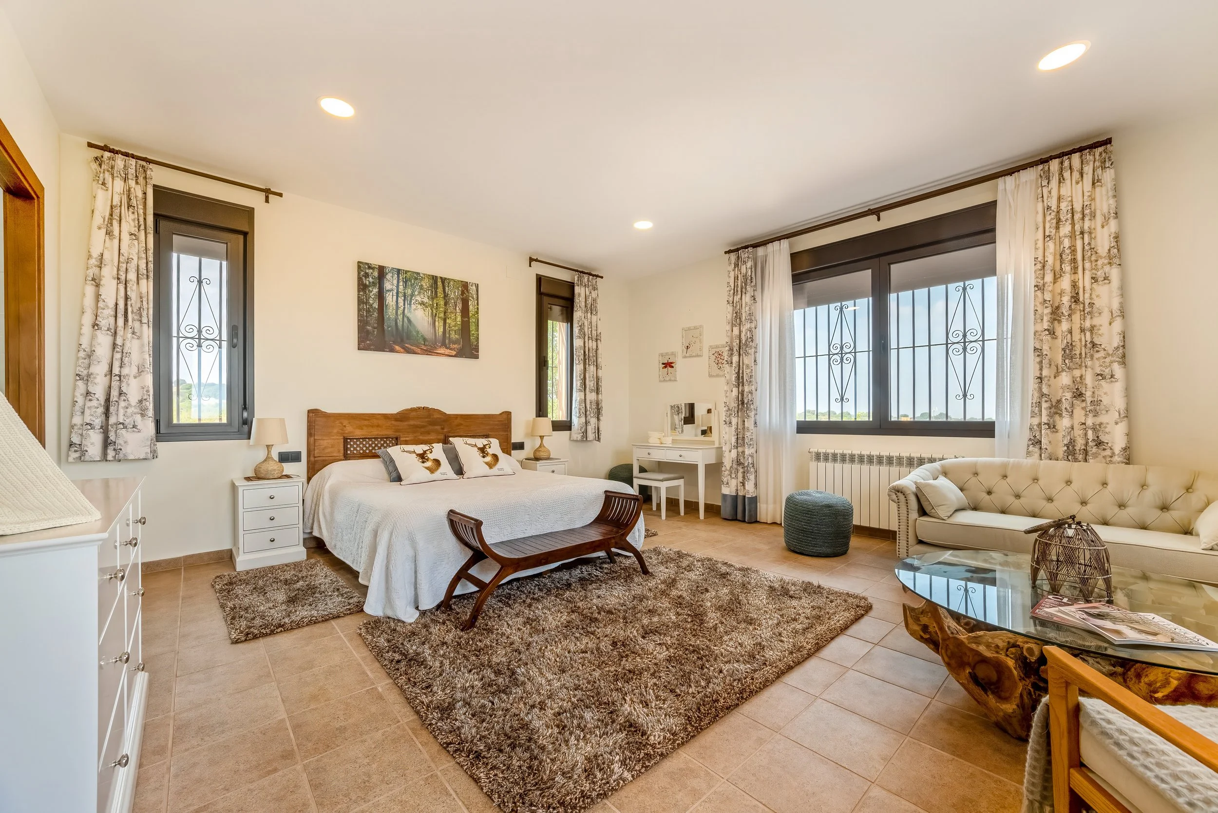 Een ruime slaapkamer met een groot bed, twee nachtkastjes, een spiegel, een bank voor het bed, een tv, een bank, een salontafel, een kast en meerdere ramen met witte gordijnen, alles in natuurlijke kleuren.