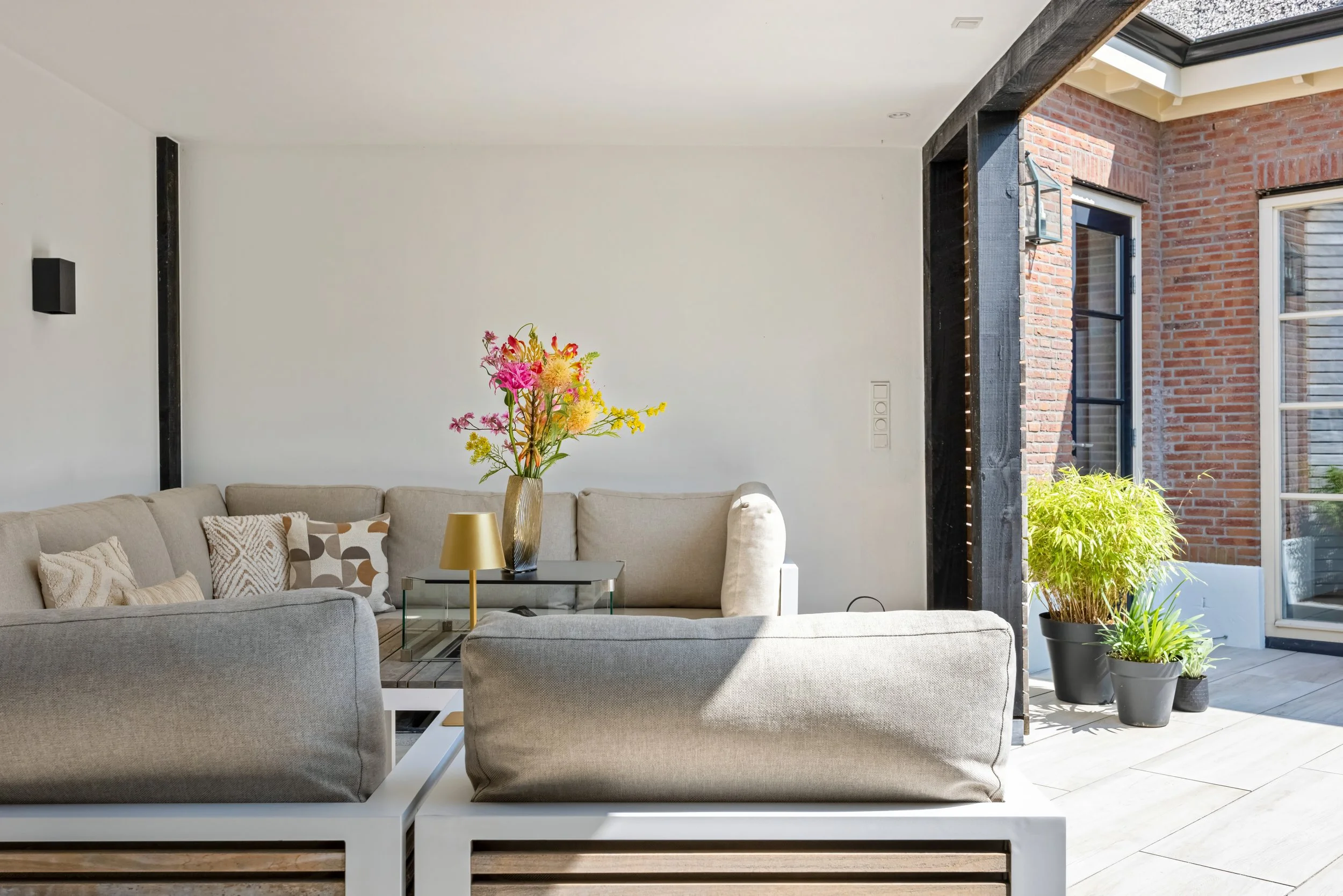 Licht interieur van een woonkamer met een beige bank, kleurrijke bloemen in een vaas op tafel, en planten in potten bij de muur met grote ramen en een open deur.