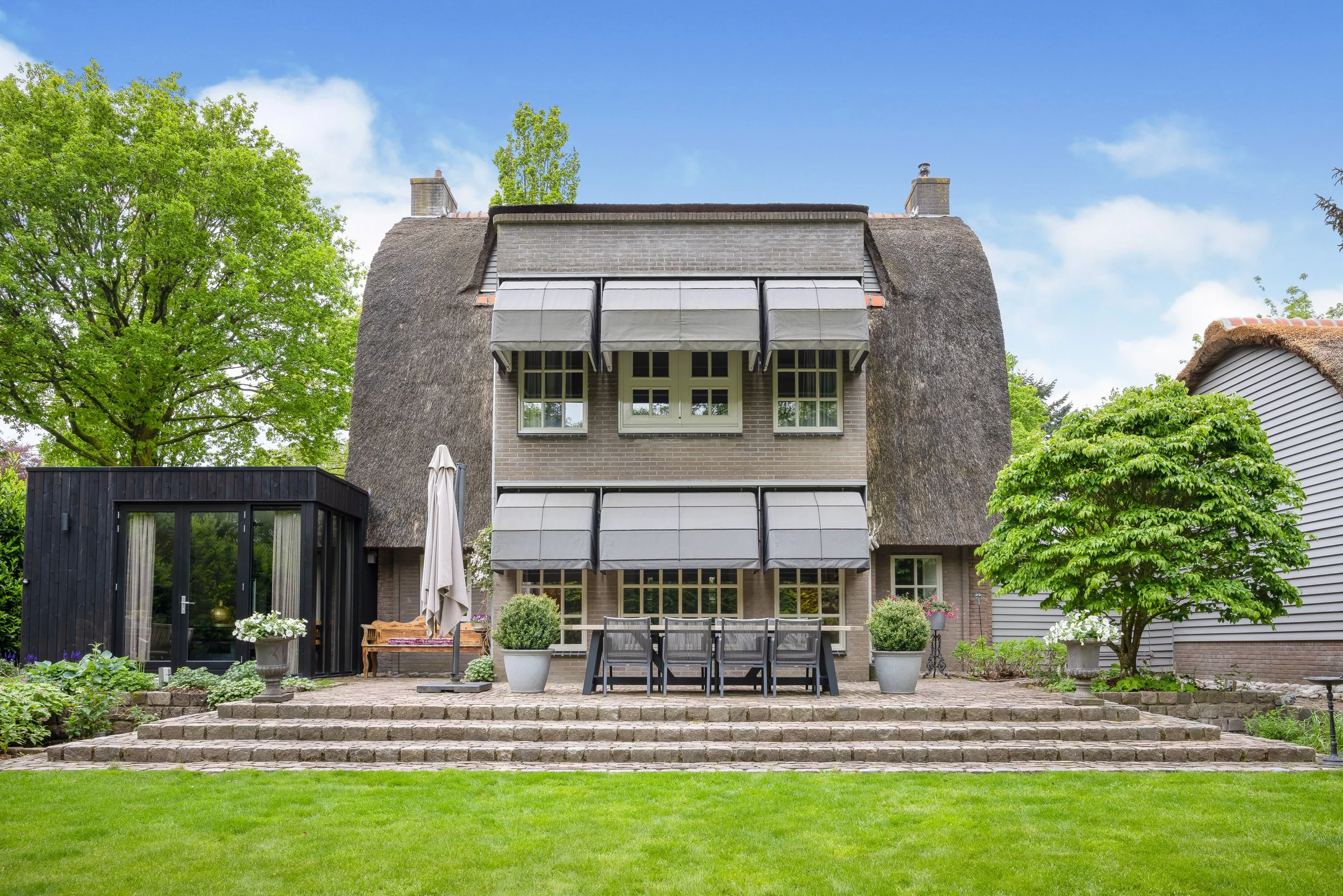Moderne woning met meerdere ramen en zonneschermen, een tuin met bomen, planten, en buitenmeubilair, inclusief een tafel, stoelen, en een parasol.