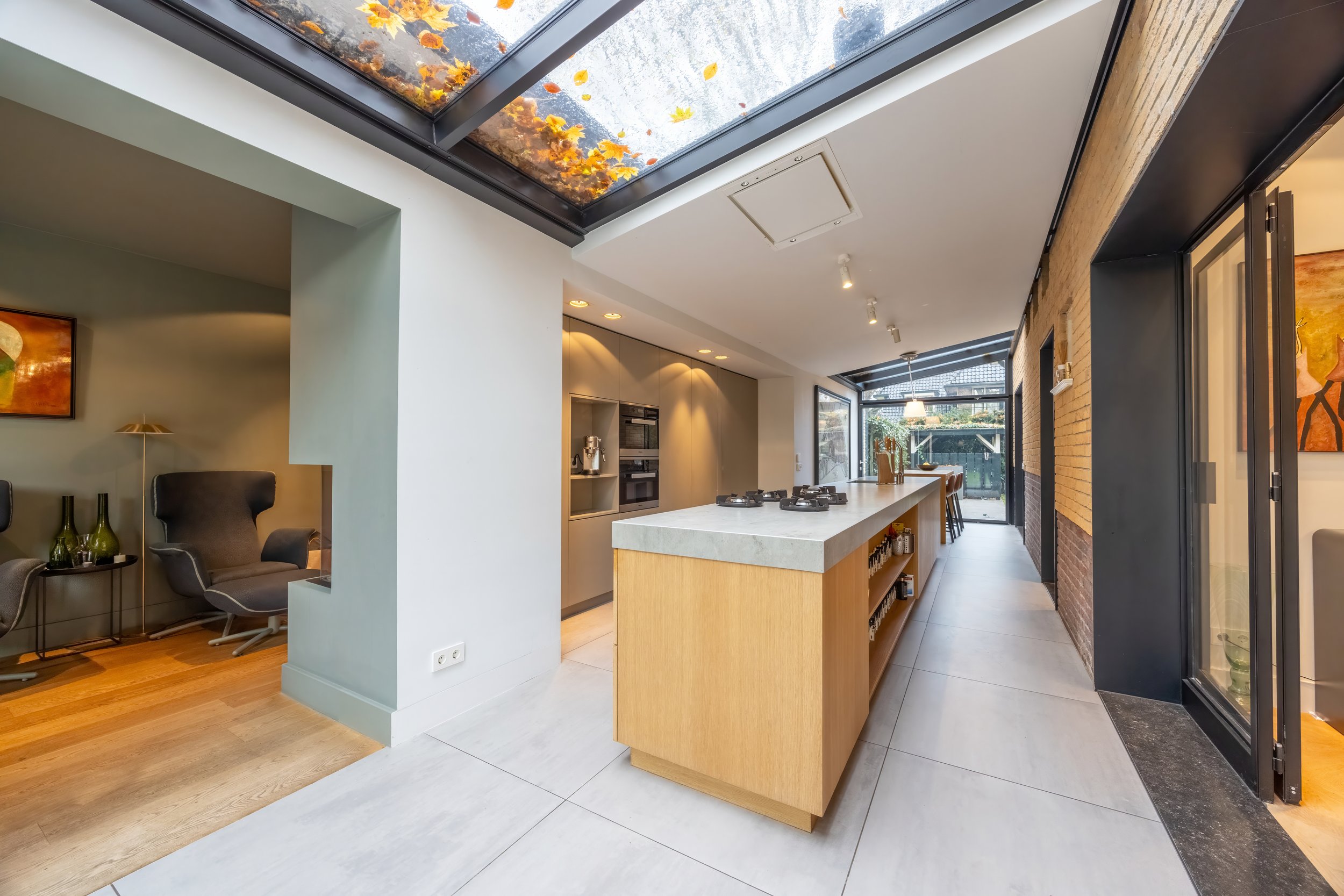 Moderne open keuken met een eiland, grote ramen en een glazen dak met bladeren in de herfst. Vakmanschap en natuur in interieur.