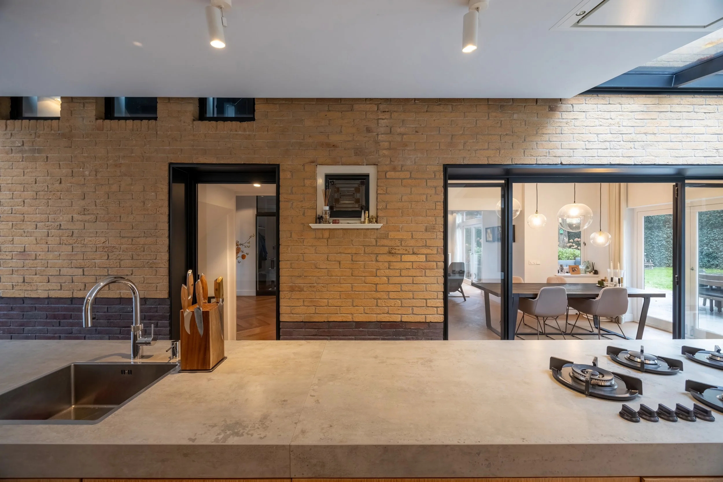 Moderne keuken met aanrecht, keukengerei, oven en een uitzicht op de eetkamer en woonkamer met grote ramen en hangende lampen.