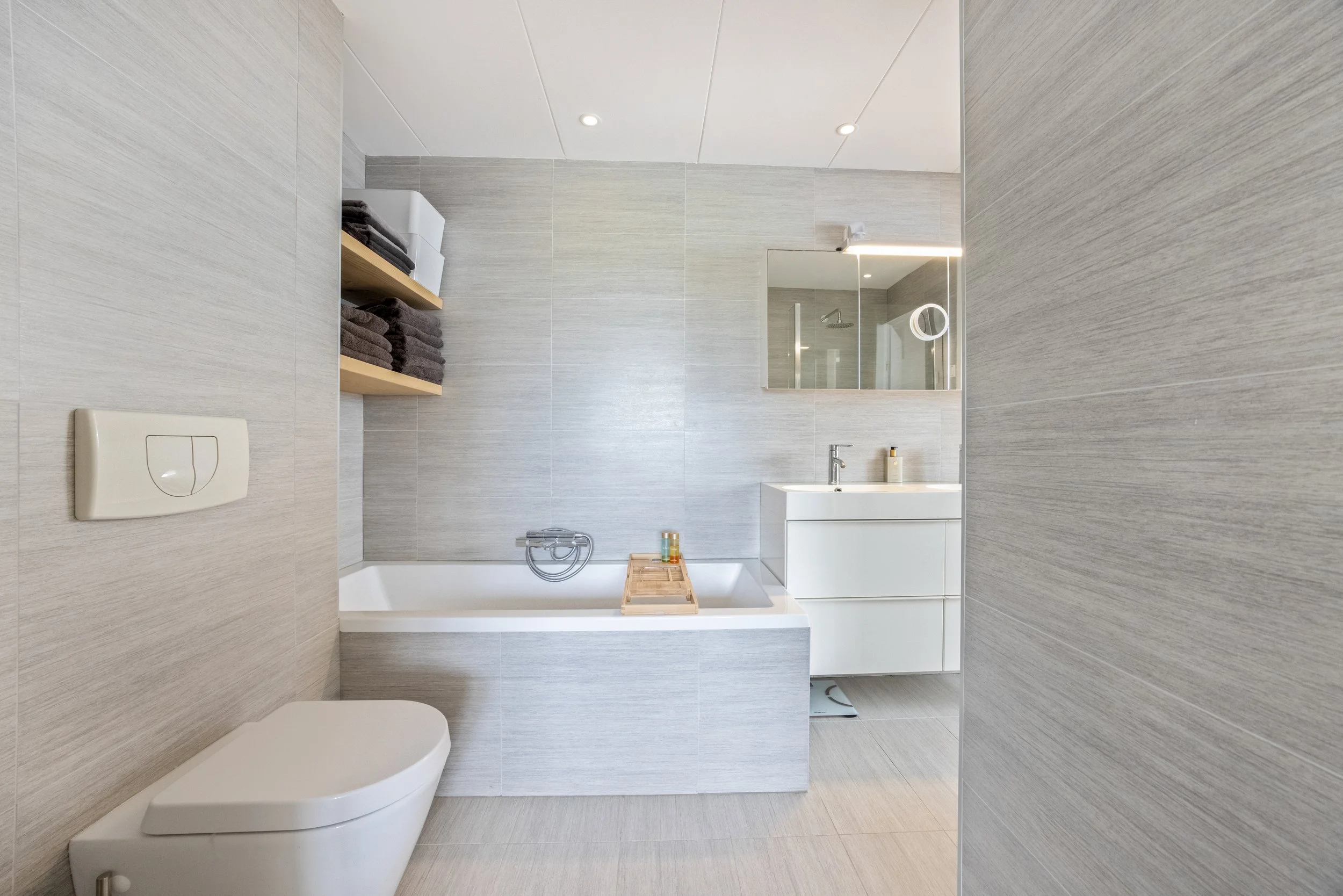 Een moderne badkamer met een ligbad, een spiegel met ingebouwd licht, een wastafel met twee lades, een toilet en opbergplanken met handdoeken.