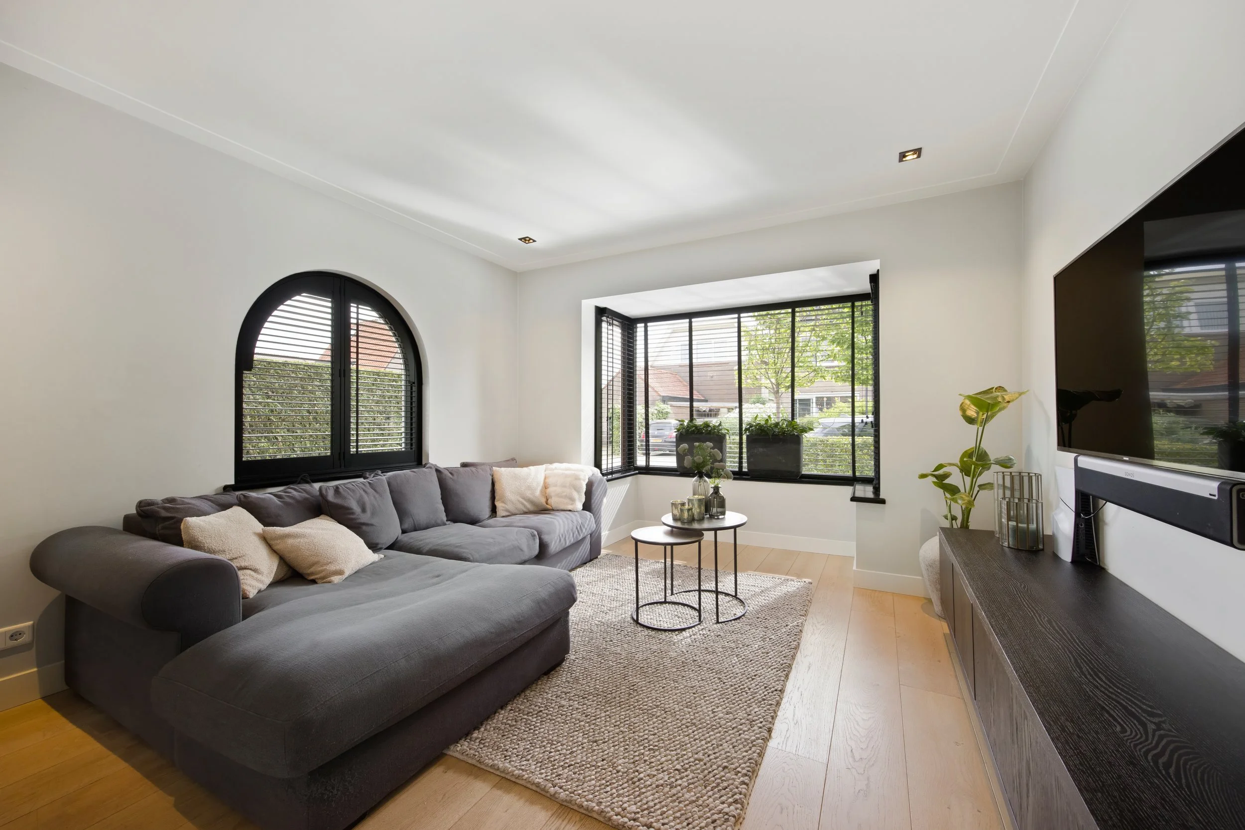 Moderne woonkamer met grote ramen, een grijze L-vormige bank met kussens, een ronde salontafel, een vloerkleed, een tv op een lage kast, planten, en een rustige decorstijl.