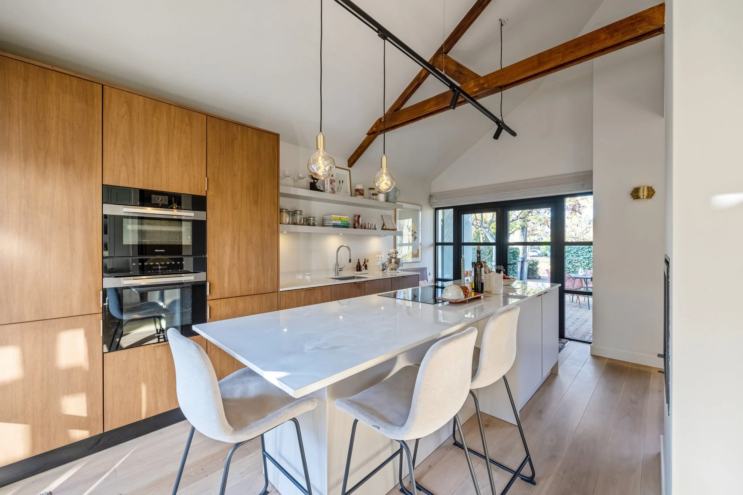 Moderne keuken met houten kasten, een wit eiland met barkrukken, open planken met servies en decoraties, grote ramen met uitzicht op tuin, houten plafondbalken.