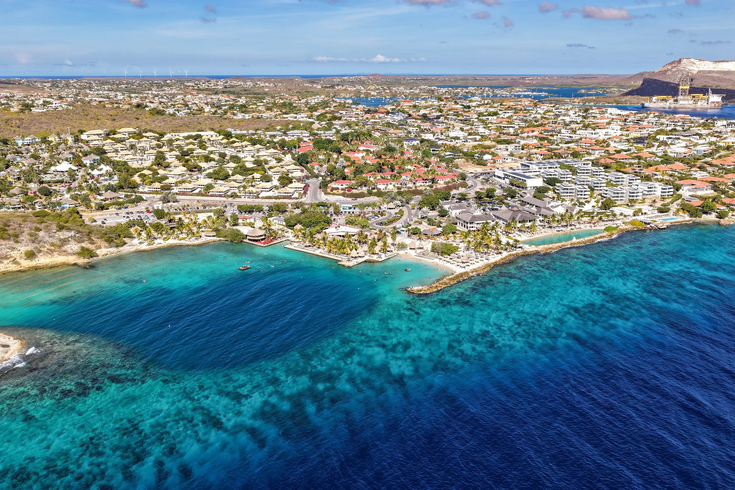 Luchtfoto van een kustplaats met helderblauw water en zandstranden, omlind door gebouwen en groenvoorzieningen, met een rotsformatie en water in de achtergrond.