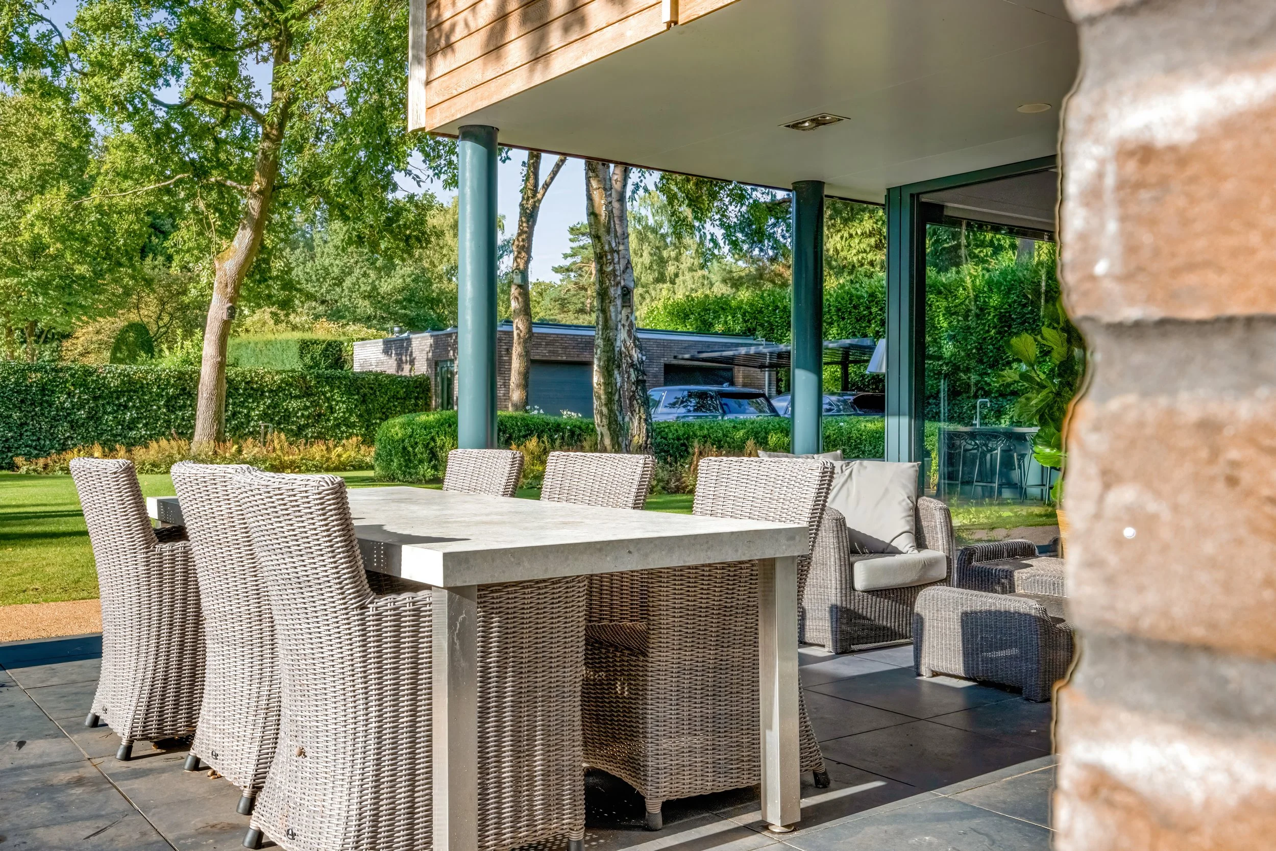 Gezellige patio met een grote witte tafel en meerdere wicker stoelen, omgeven door groene bomen en struiken, met een modern huis op de achtergrond.