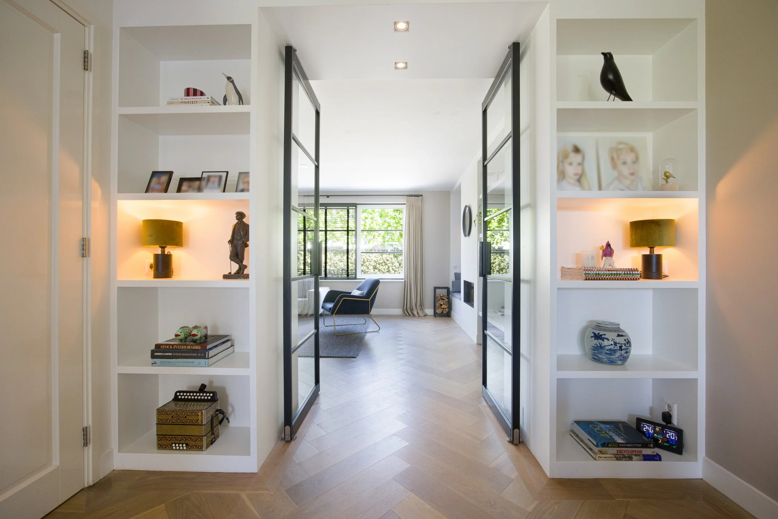 Foto van een lichte woonkamer met grote ramen, een blauwe stoel, en een open haard. Aan weerszijden van de deur naar de woonkamer bevinden zich ingebouwde witte planken met decoratieve objecten en lampen.