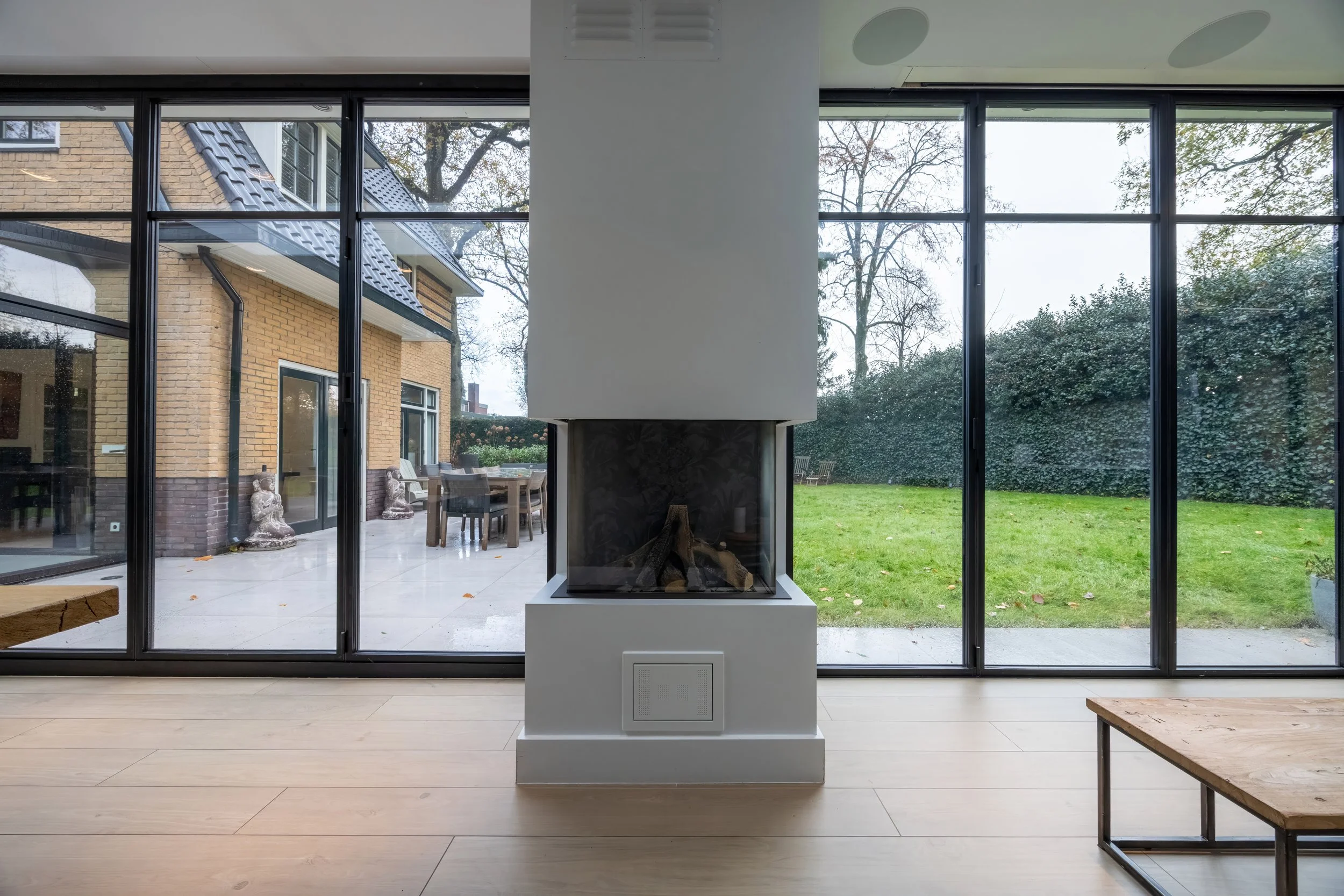 Binnenkant van een huis met grote glazen schuifdeuren die uitkijken op een tuin met gras en bomen, centrale klommutz fireplace met houtblokken binnen, houten vloer en een salontafel aan de rechterkant.