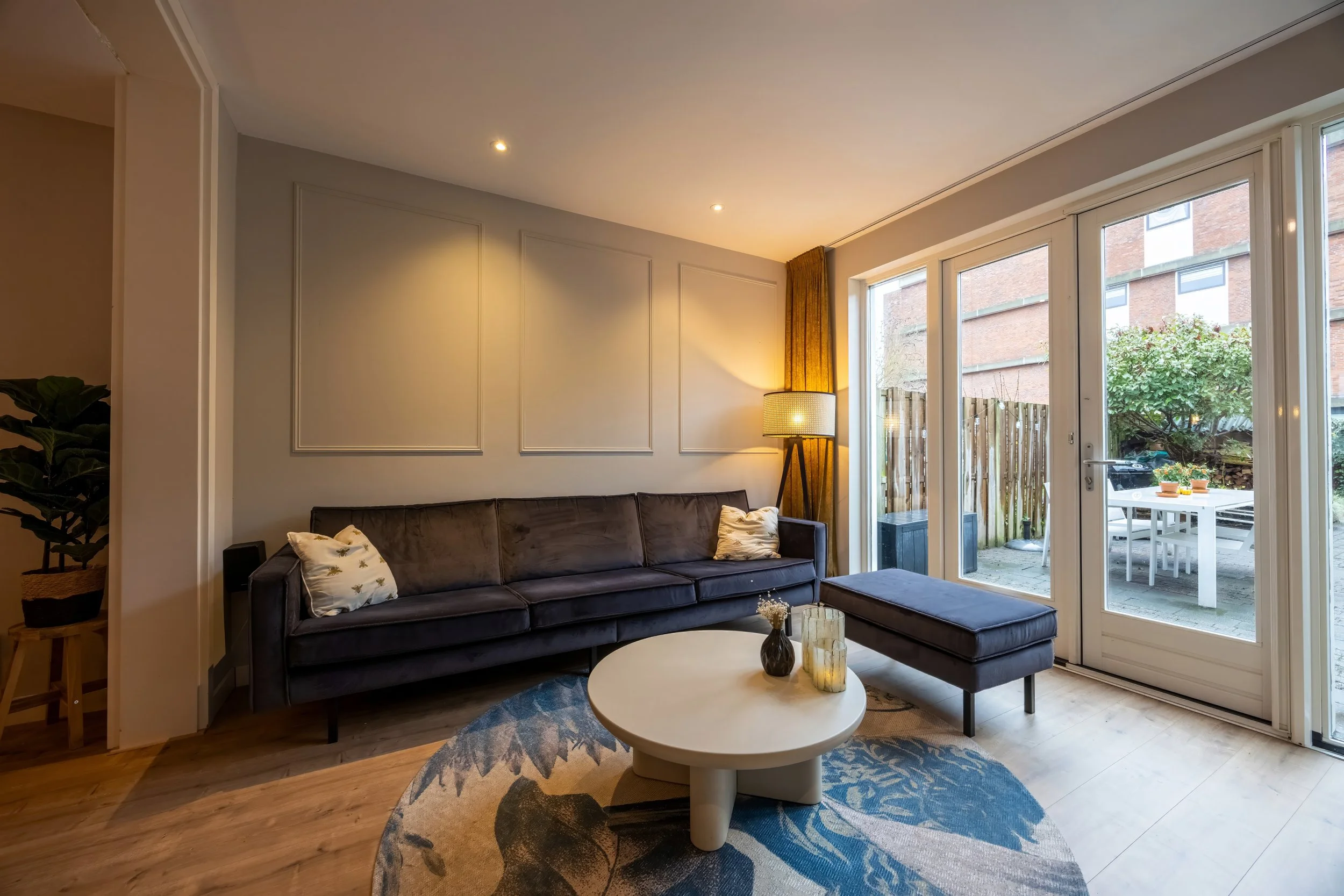 Woonkamer met een donkerbruine bank, een witte ronde salontafel met vazen, een vloerkleed met een bladpatroon, grote glazen deuren naar een tuin met buitentafels en stoelen, en een grote vloer-lamp naast de deur.