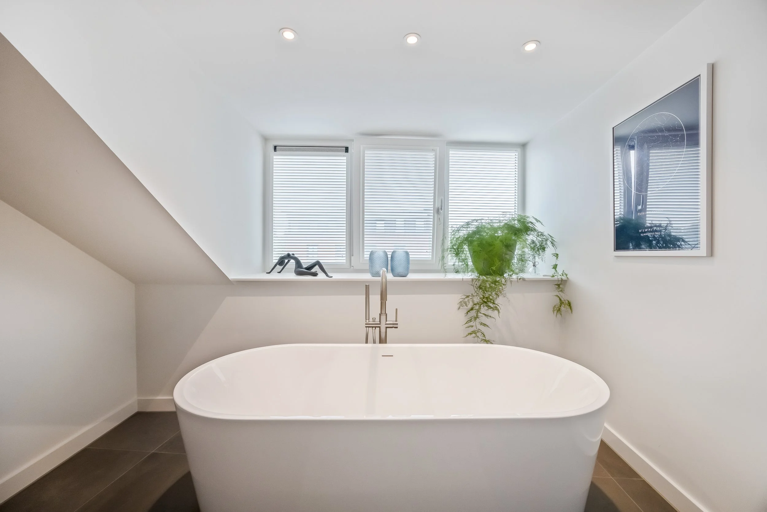 Badkamer met vrijstaand bad, grote raampartijen met jaloezieën, planten en muurkunst.