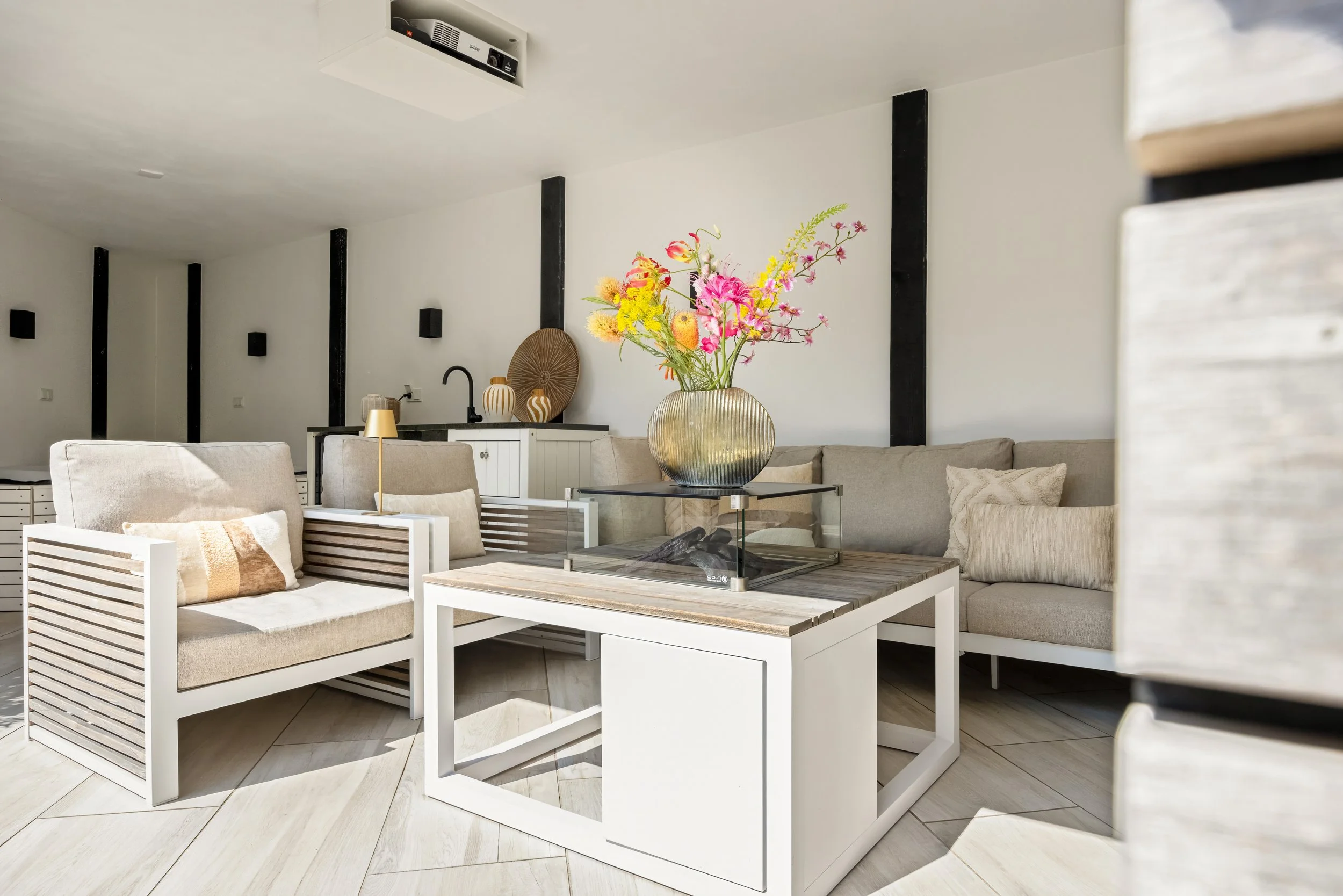 Woonkamer met beige banken, witte salontafel met glazen vaas met bloemen, open keuken op achtergrond.