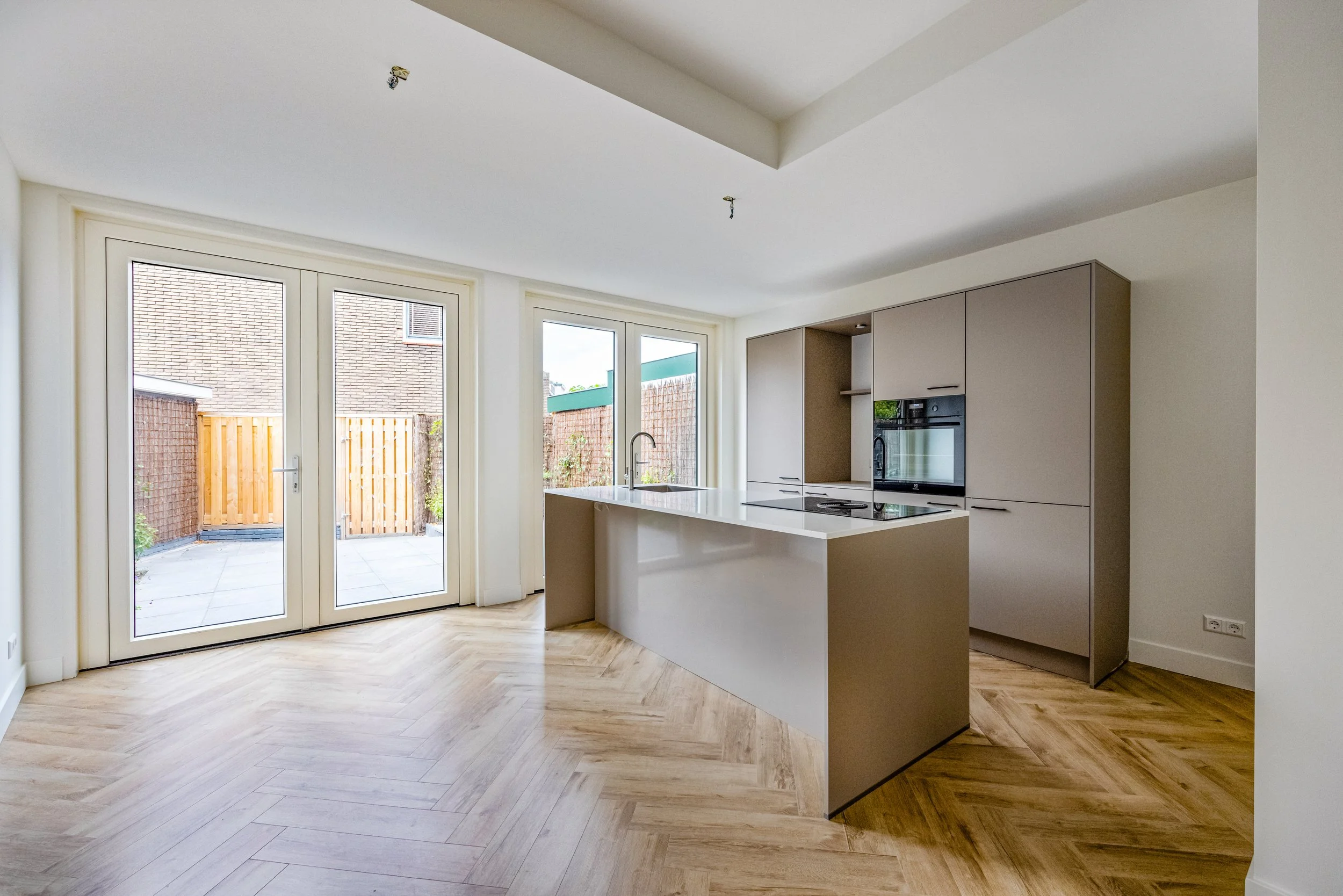 Moderne, lege keuken met grote glazen deuren naar de patio, licht houten vloeren, beige kasten, eiland met oven en kookplaat.