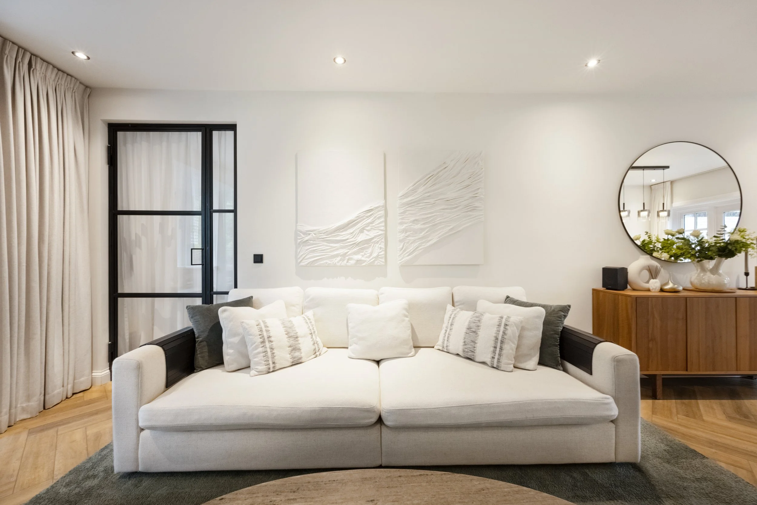 Woonkamer met een witte bank met meerdere kussens, houten vloer, ronde spiegel, plant, en moderne decoraties