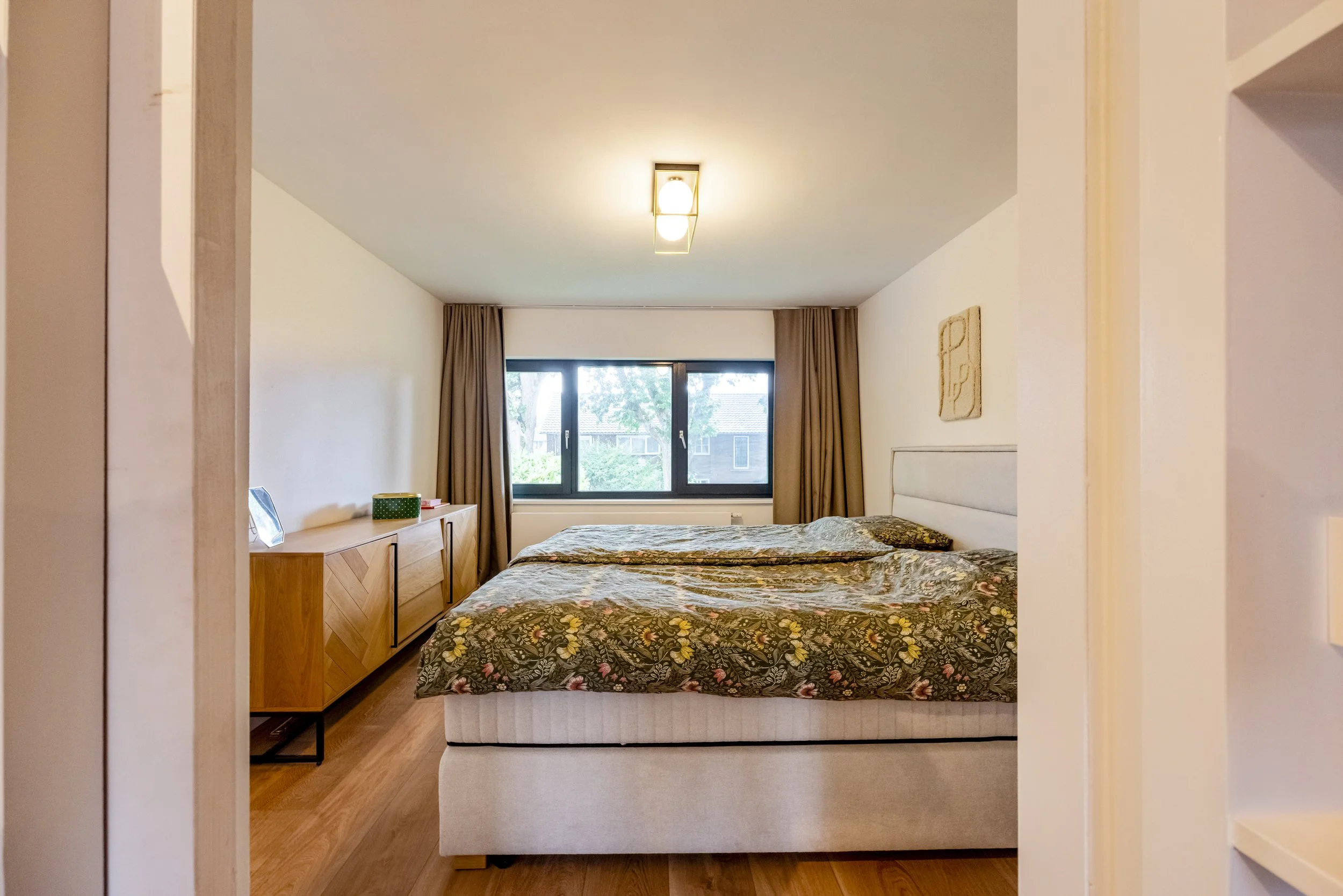 Woonkamer met groot bed, houten kast, raam met uitzicht op bomen, en beige muren.