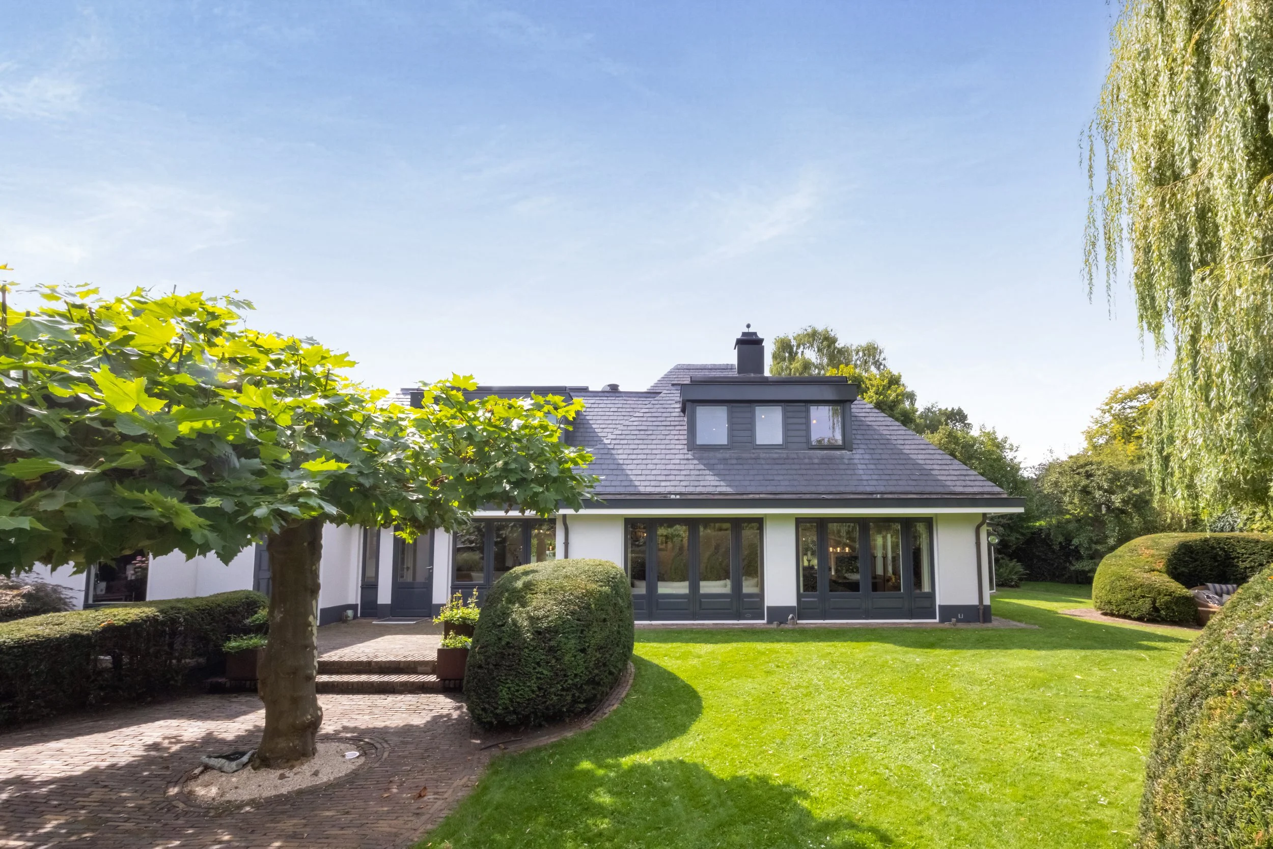 Witte huis met donkere ramen en een schoorsteen, omgeven door een groene tuin met gras, bomen en struiken, onder een blauwe lucht.