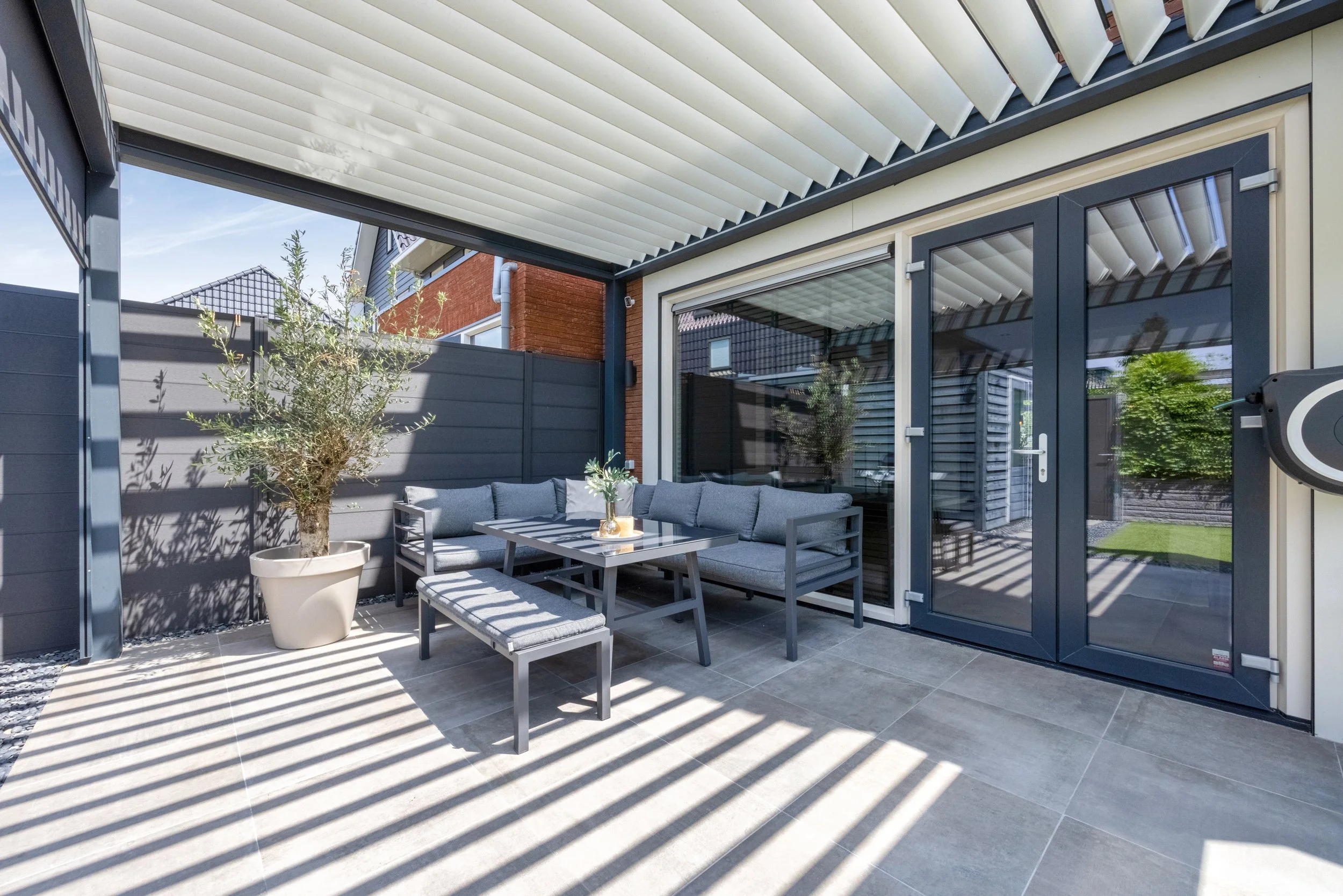 Een moderne buitenpatio met een overkapping, een hoekbank met kussens, een tafel met een vaas en een boom in een grote pot, omringd door een hoge schutting en glazen deuren.