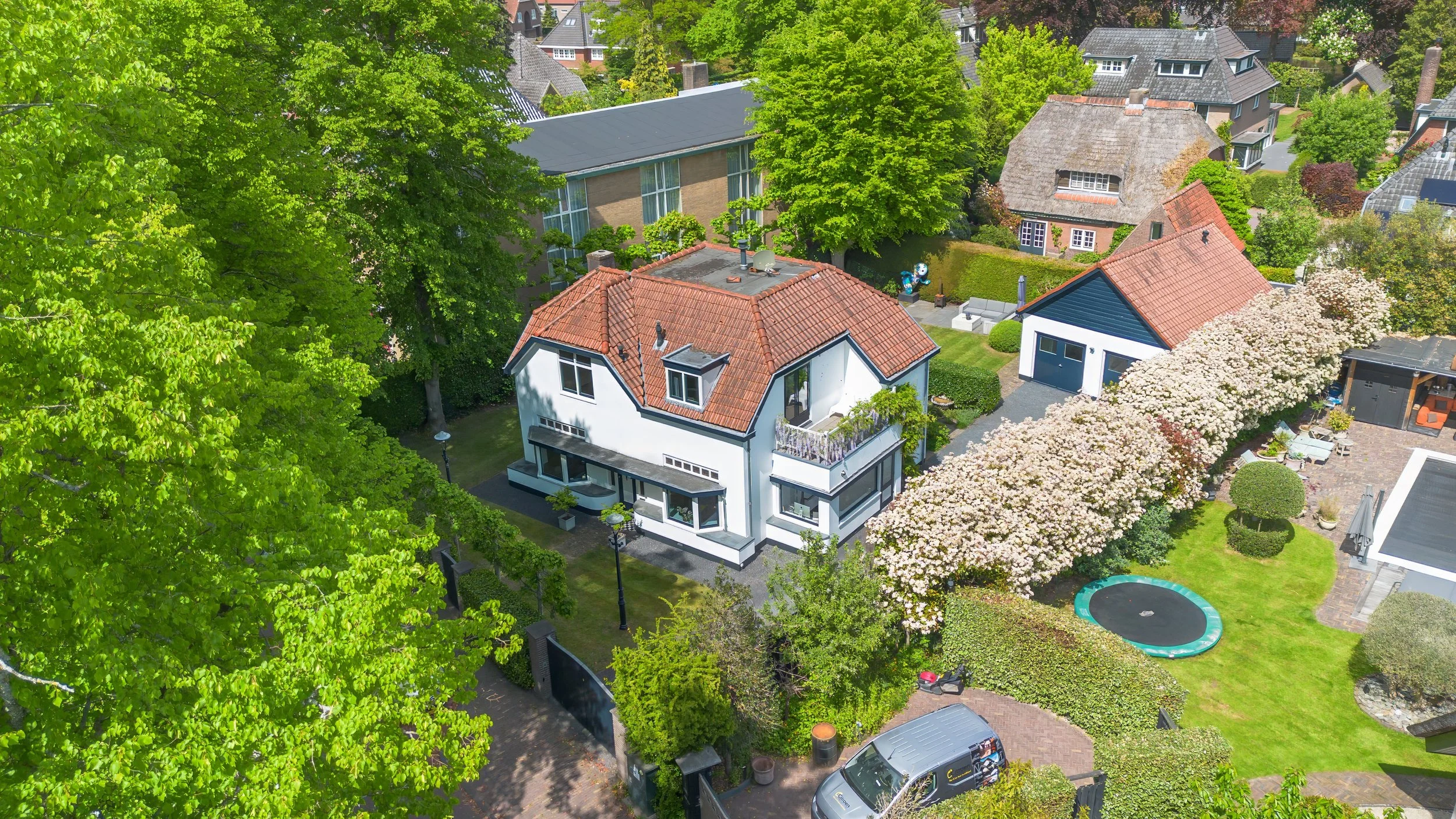 Uitzicht van bovenaf op een witte woning met een rode dak, omgeven door groene bomen en een tuin met een trampoline en een parasol.