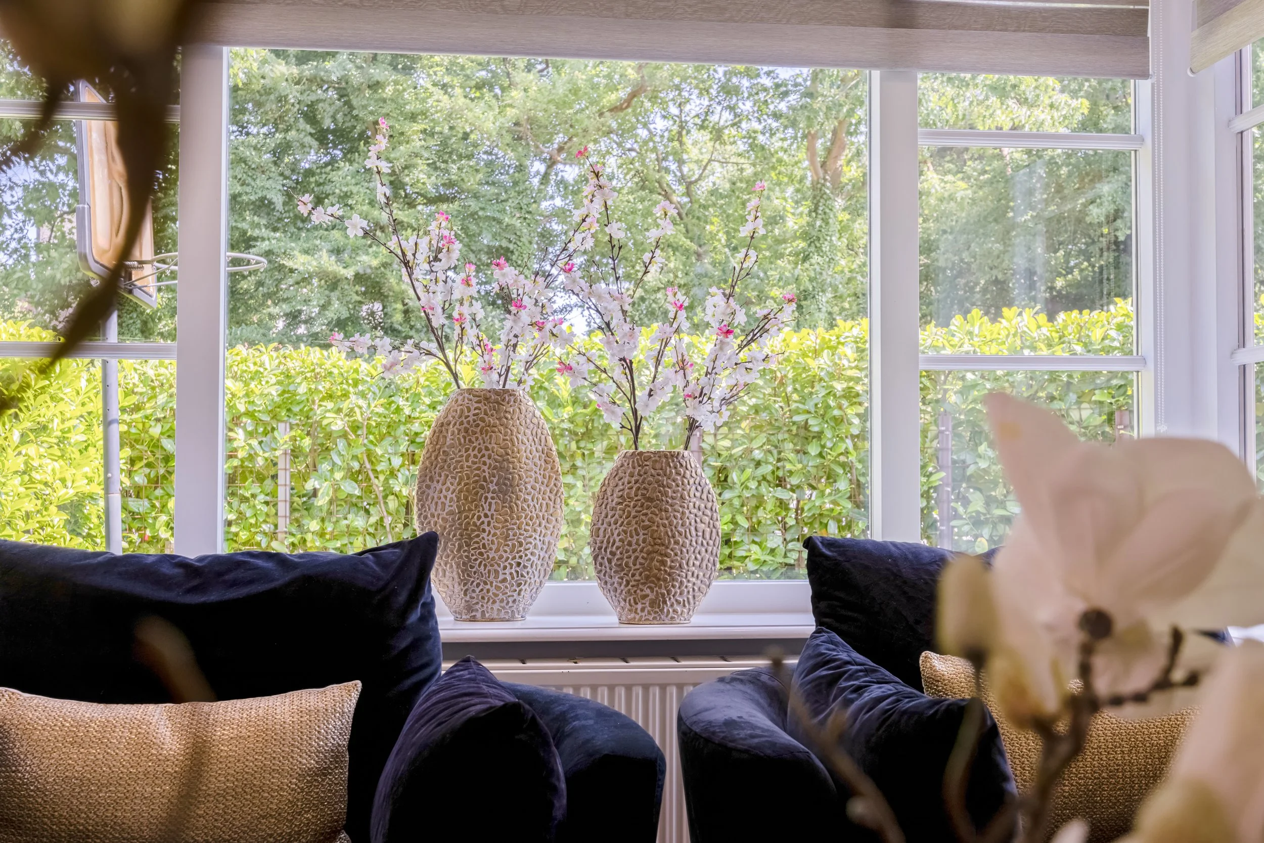 Interieur van een woonkamer met twee beige vazen met bloemen op de vensterbank, omringd door zwarte en goudkleurige kussens en een grote witte orchidee in de voorgrond, met een uitzicht op groene bladeren buiten.