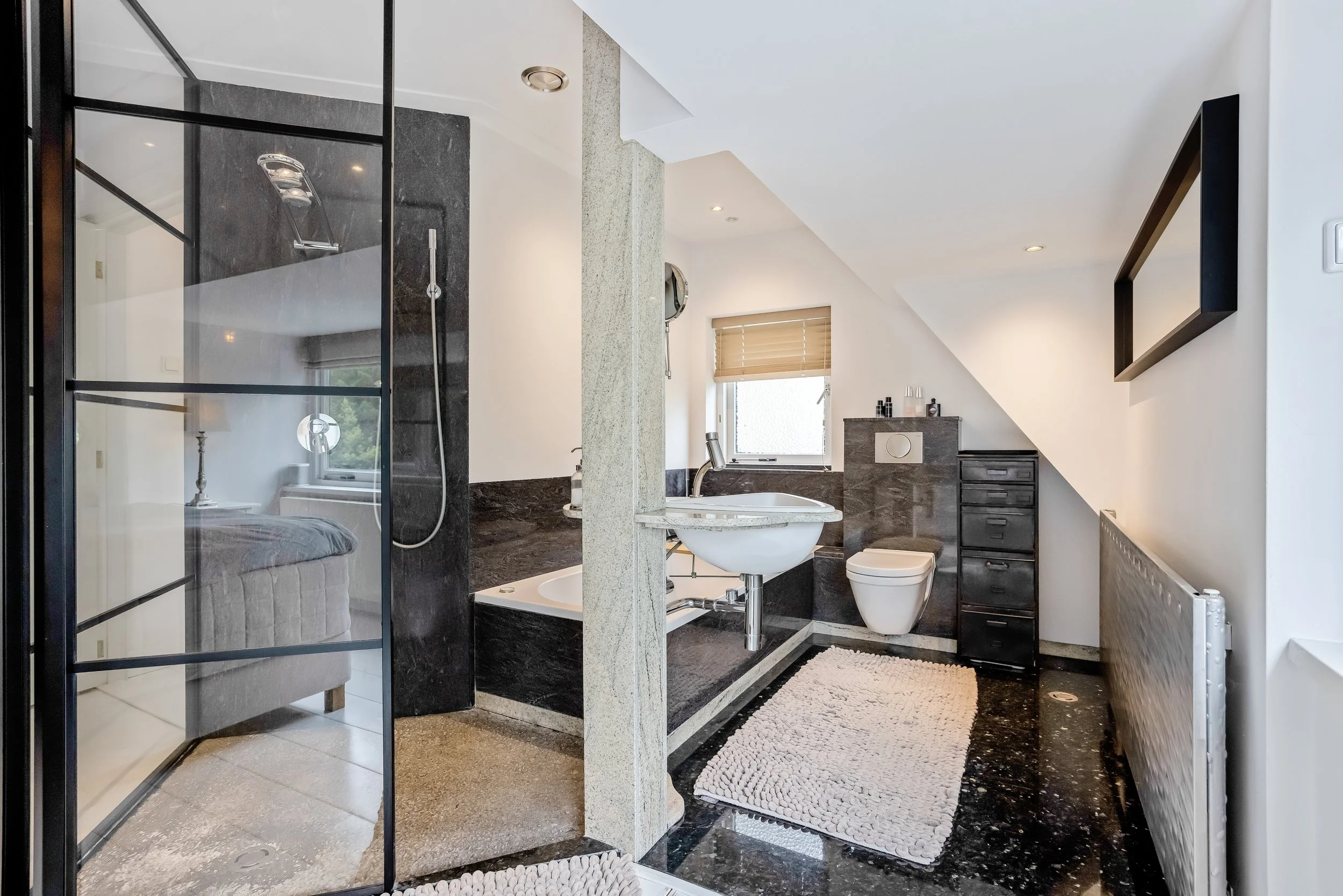 Een moderne badkamer met een inloopdouche, een vrijstaande badkuip, toilet en zwarte meubelstukken, inclusief handdoekradiator, op een zwart granieten vloer.