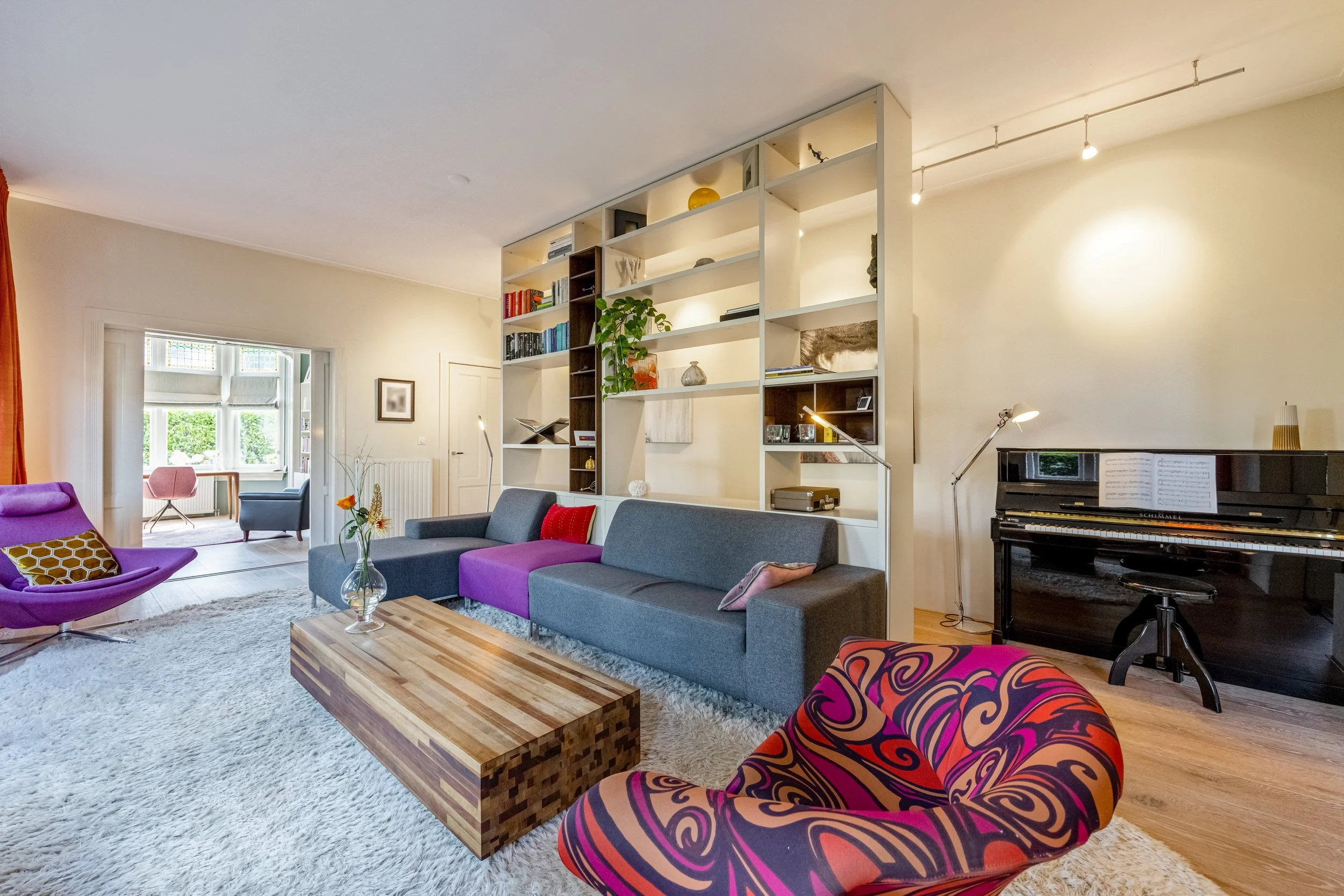 Woonkamer met kleurrijke kuipstoelen, een grijze bank met paar kussens, een boekenplank afscheiding, een vleugel en een openzitje bij het raam