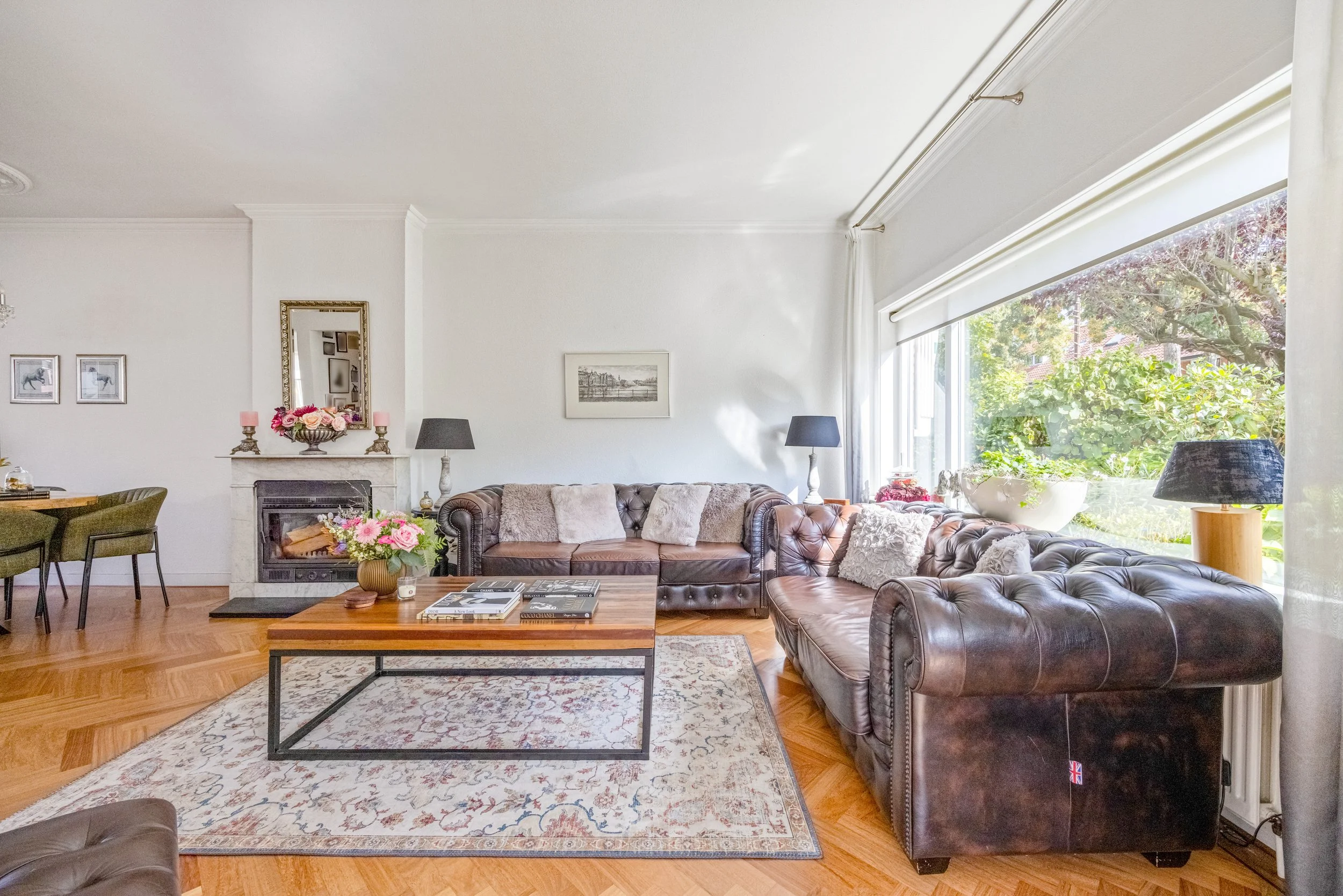 Luxe woonkamer met leren bank, salontafel, weblogs, open haard, decoraties en grote ramen met uitzicht op een tuin.