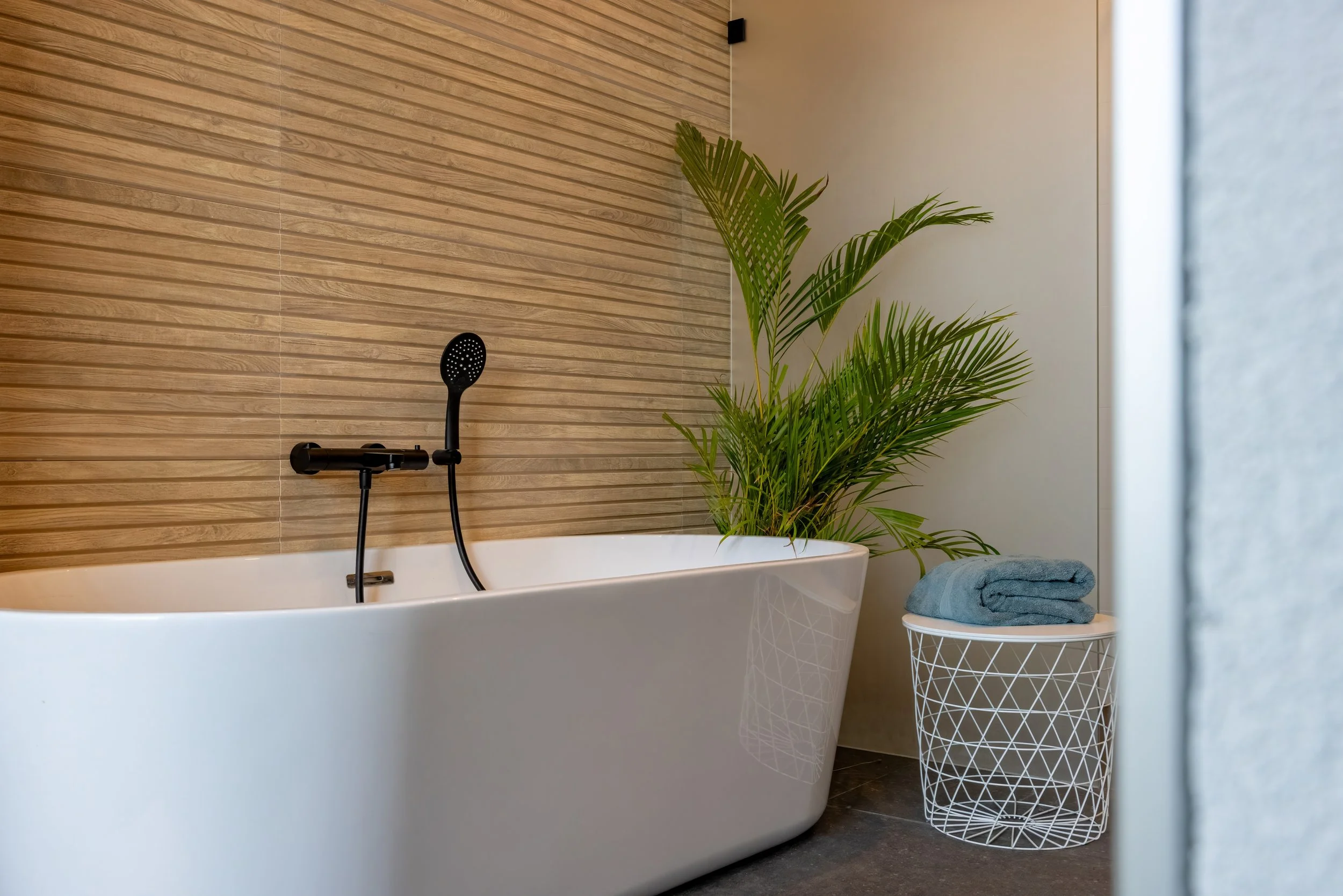Een witte vrijstaand bad met een zwarte handdouche, een groen planten in de hoek en een mand met opgerolde handdoeken naast de muur.