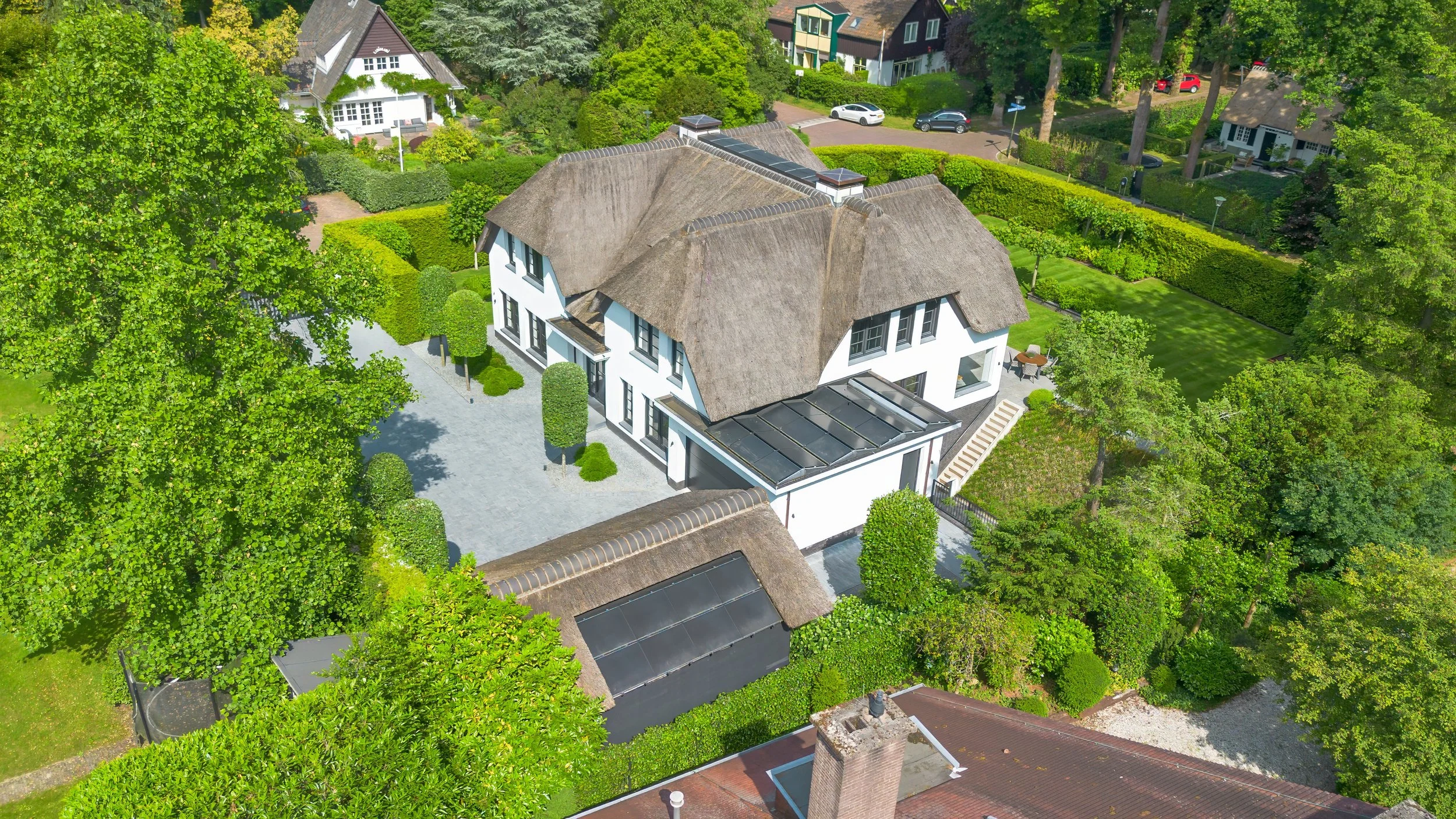 Luchtfoto van een wit huis met datagedeelte en een rieten dak, omgeven door een tuin met diverse bomen, struiken en een terras.