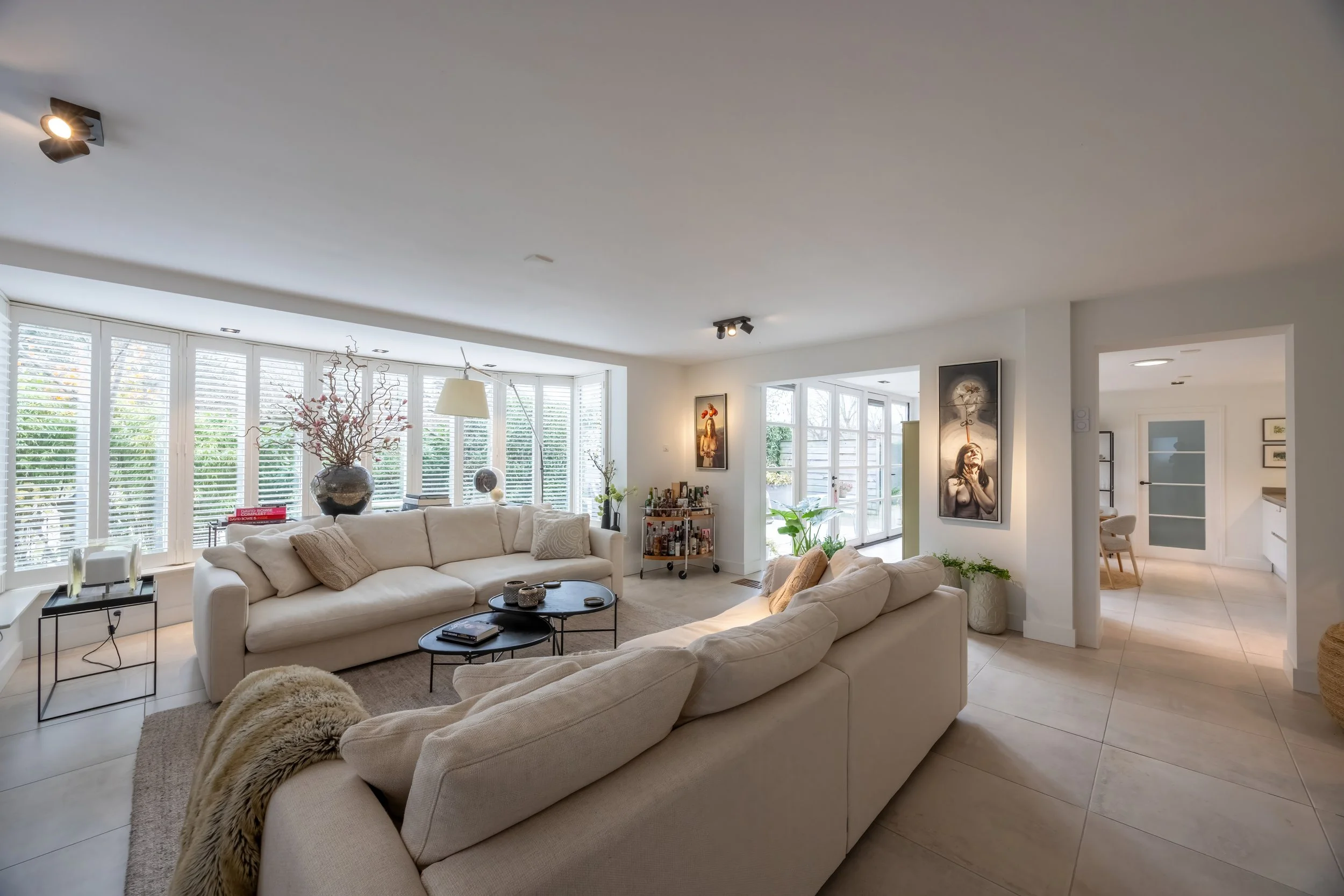 Lichte woonkamer met grote ramen, beige banken, decoratieve kunst en planten.