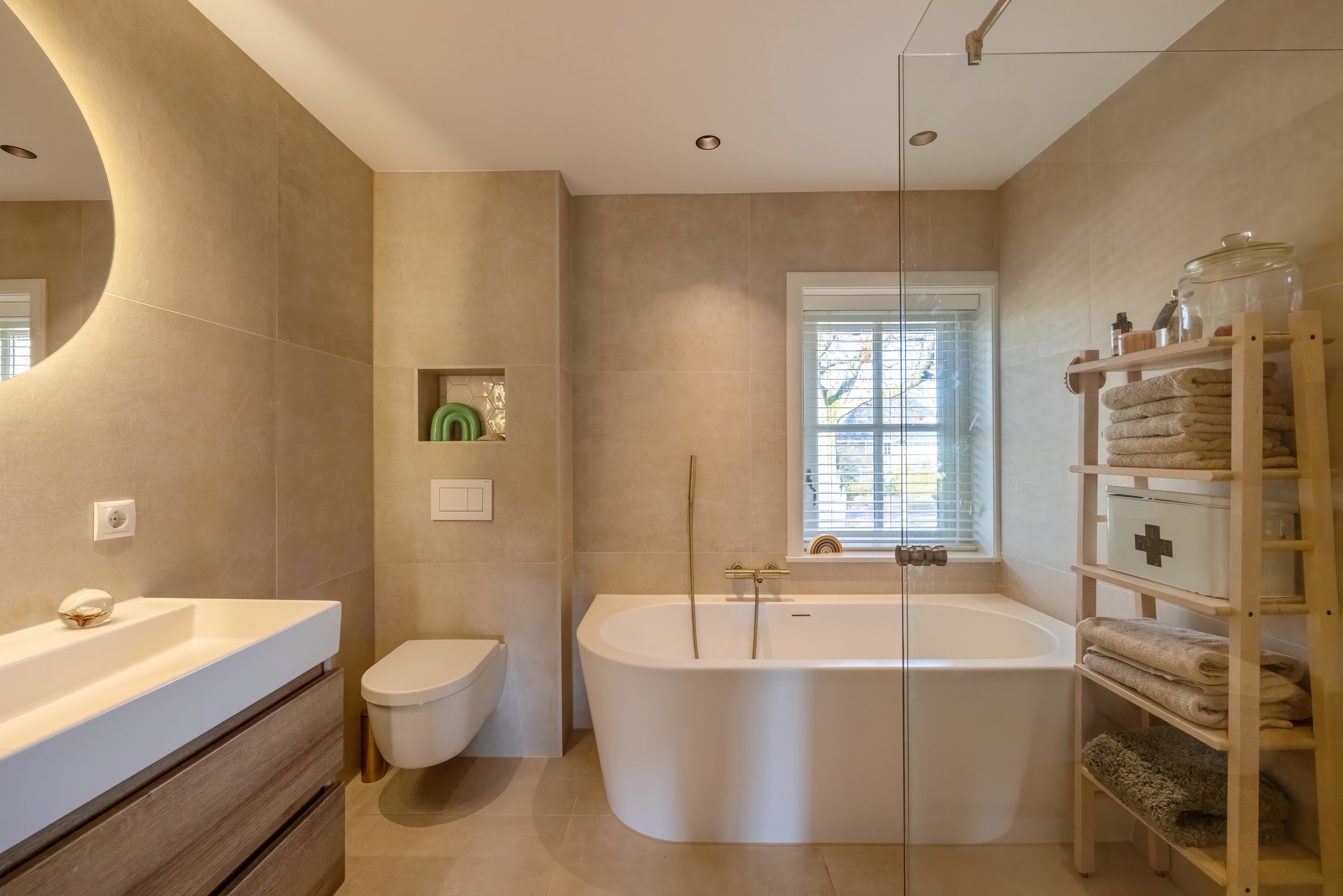 Moderne badkamer met ligbad, toilet, wastafel en opbergrek met handdoeken in natuurlijke kleuren.