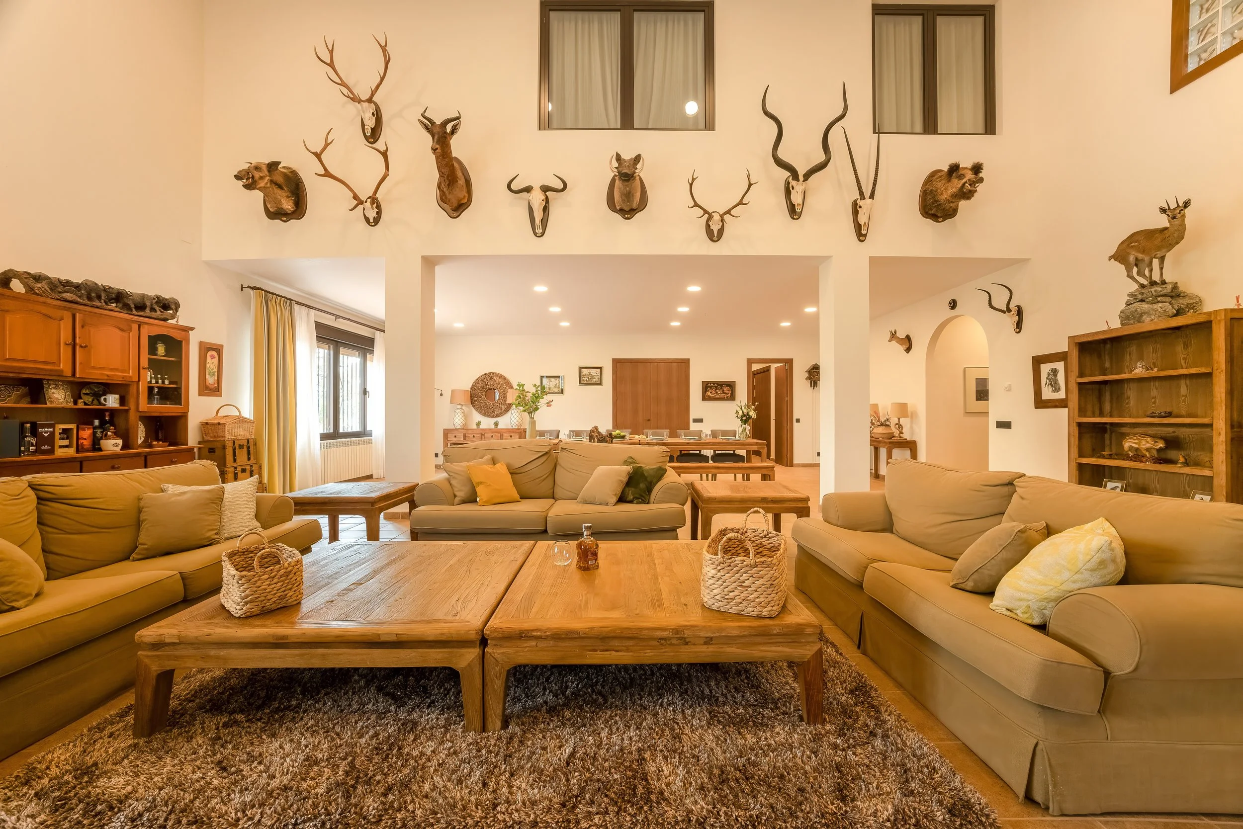 Een gezellige woonkamer met beige banken, houten salontafels, een donkerbruine tapijt en muurdecoraties van dierenhoofden en schedels.