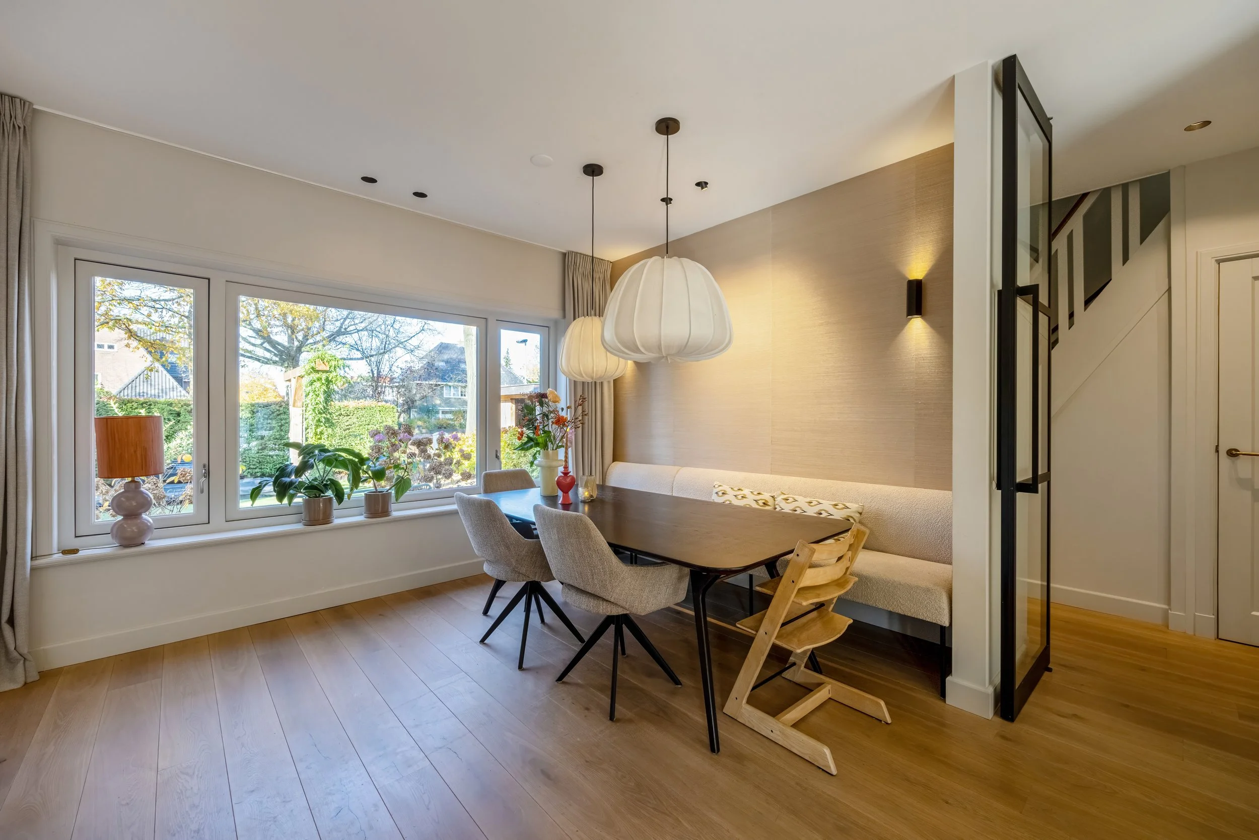 Eetkamer met grote raam, hanglampen, planten en houten vloer.