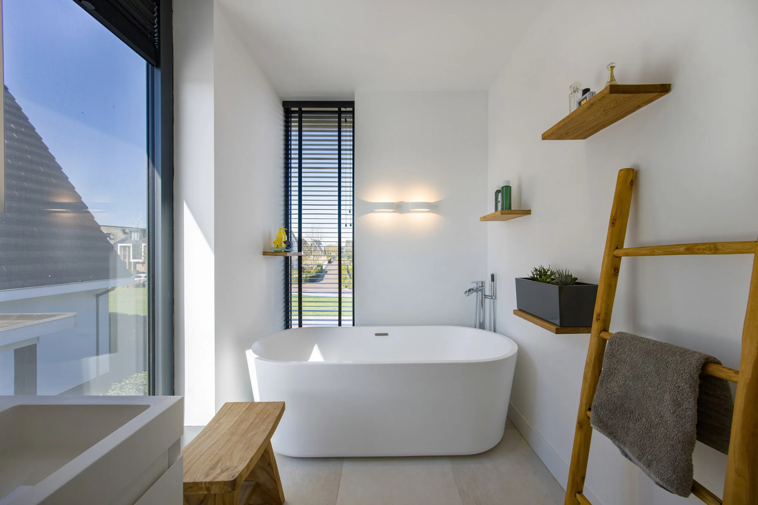 Moderne badkamer met een vrijstaand bad, grote ramen, houten schappen en een ladder met een handdoek.