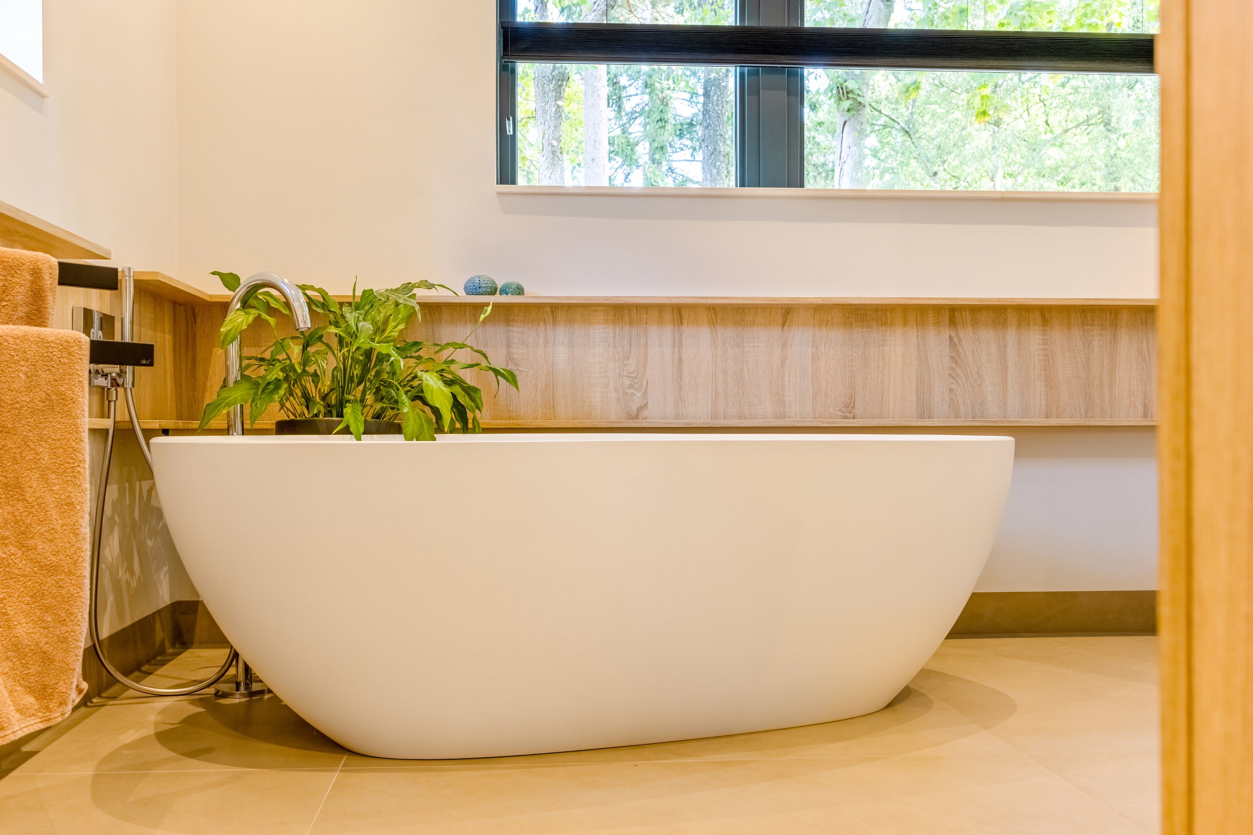 Moderne vrijstaande witte badkuip in een badkamer met grote raam, houten wandbescherming en een groene plant op de rand.