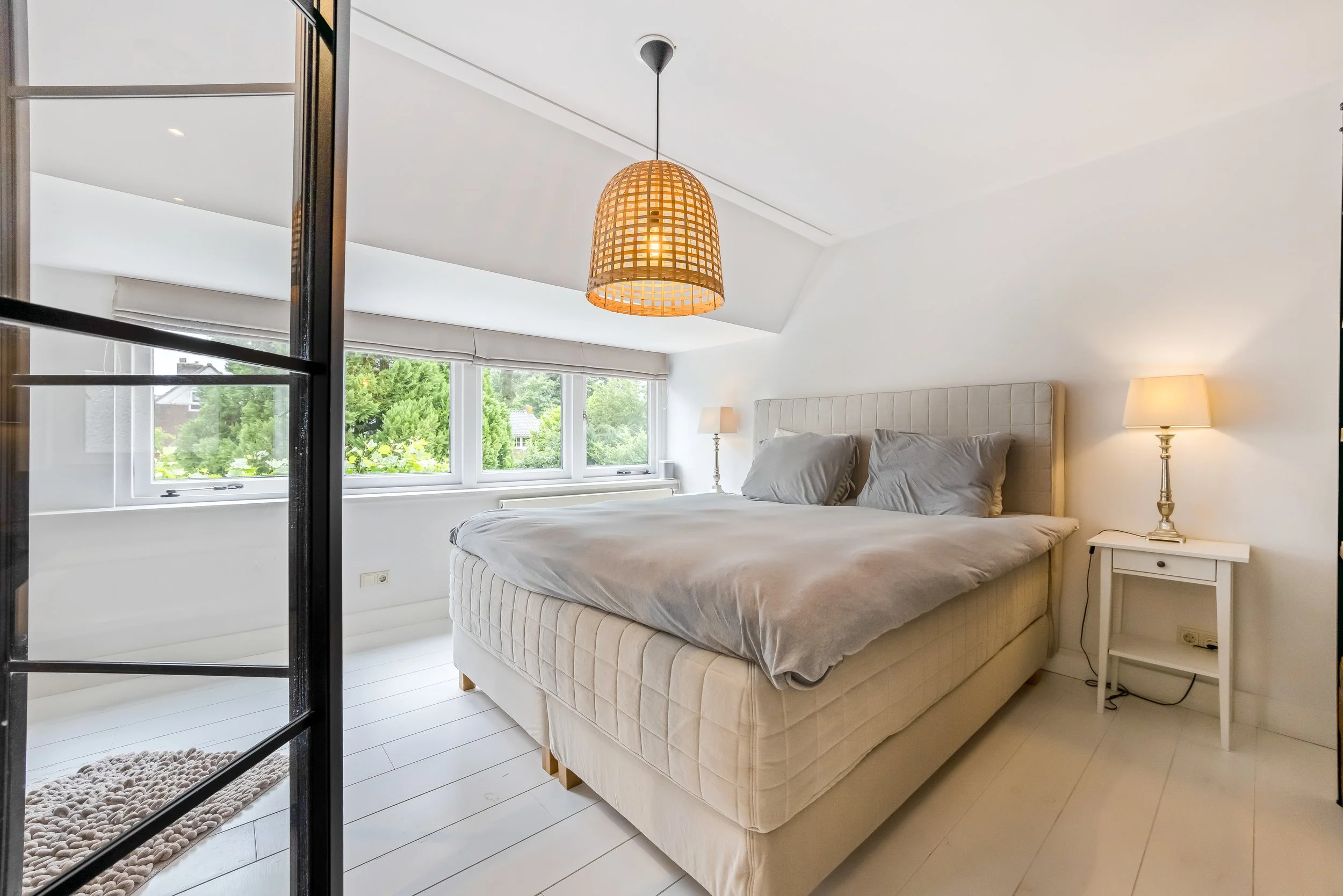 Slaapkamer met wit bed, grijze lakens, witte nachtkastjes en lampen, grote ramen met uitzicht op groen tuin, witte muren en houten vloer.