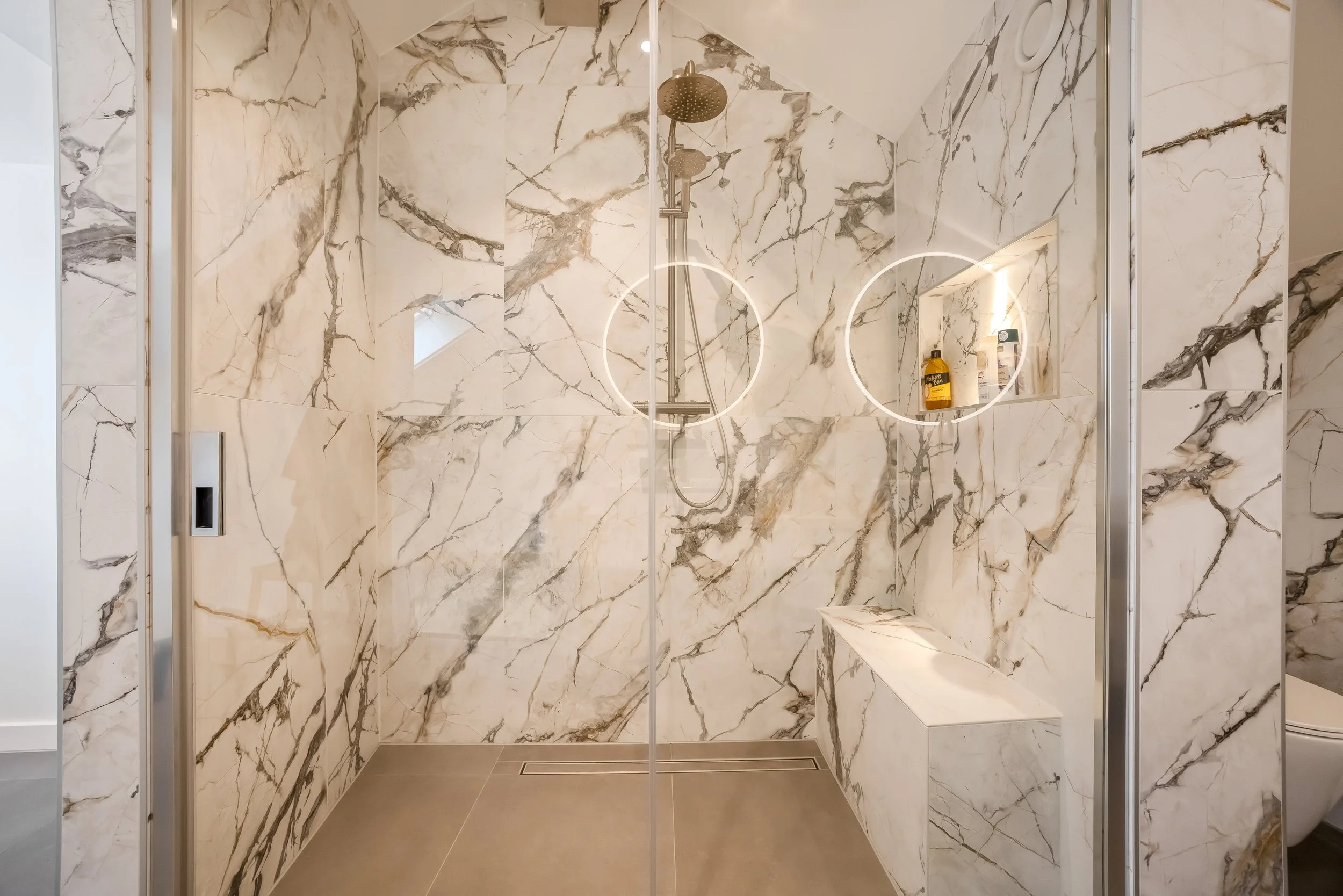 Moderne inloopdouche met marmeren wanden en een zitbank, voorzien van ronde LED-verlichting en een metalen douchekop.