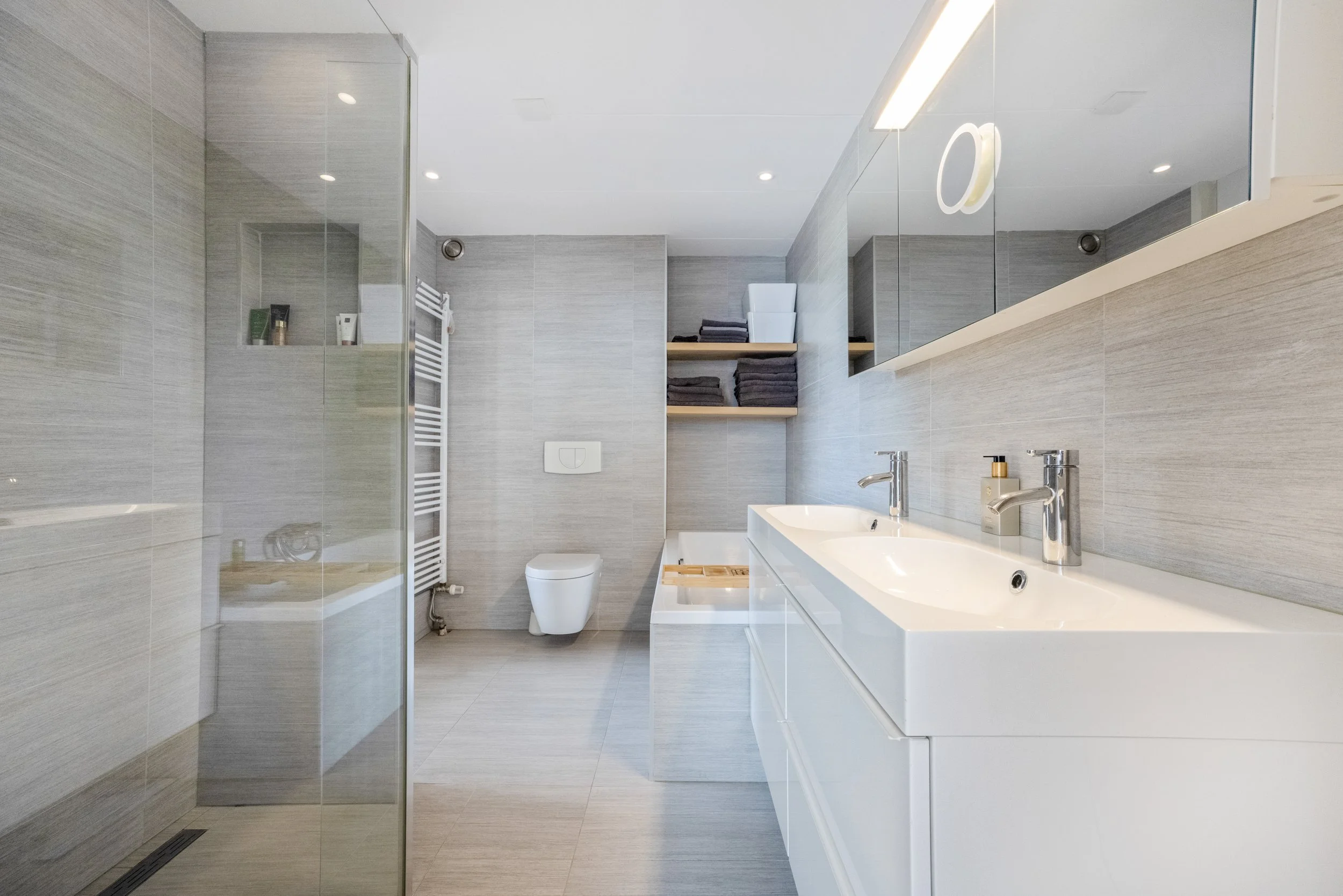 Moderne badkamer met grote spiegel, dubbele wastafel, toilet en inloopdouche met glazen wand. Opbergruimte met handdoeken en toiletries.