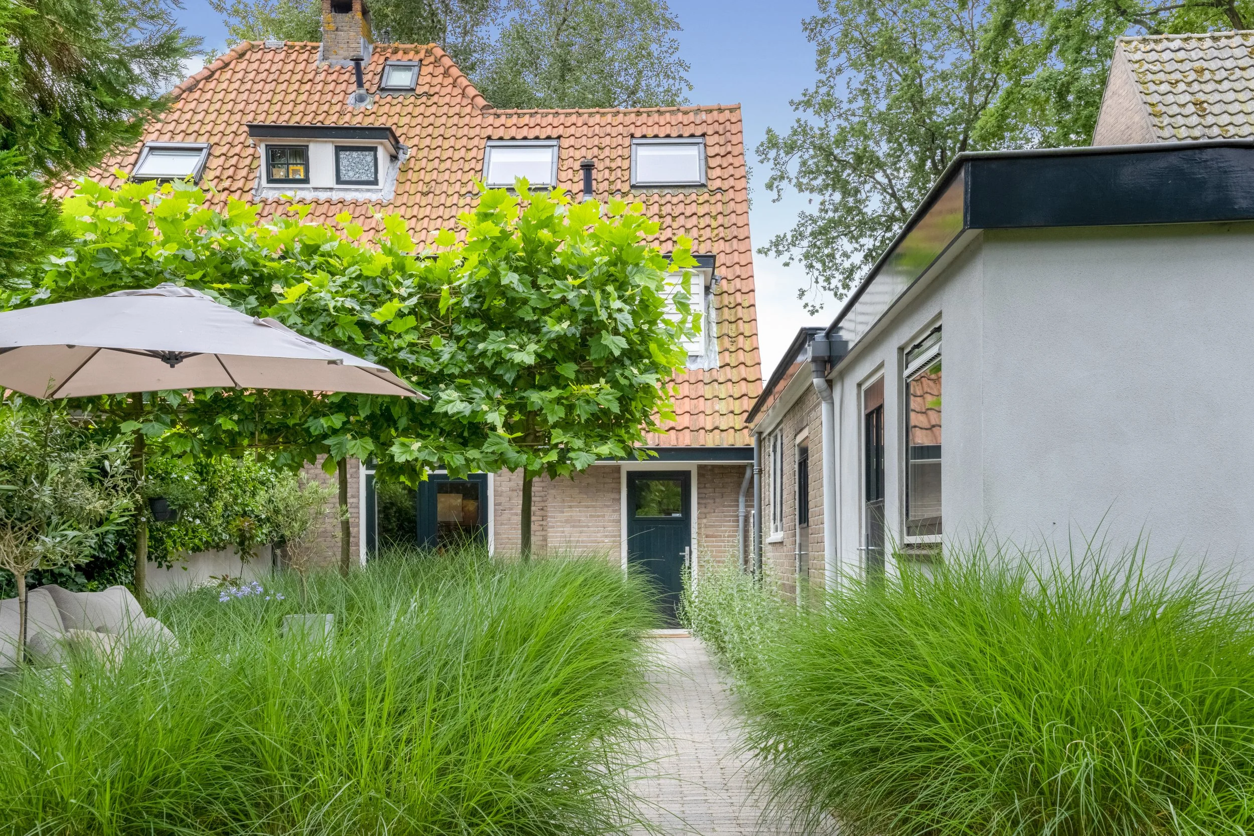 Tuin met gras, planten, een parasol en een pad voor een huis met een rode daken en meerdere ramen