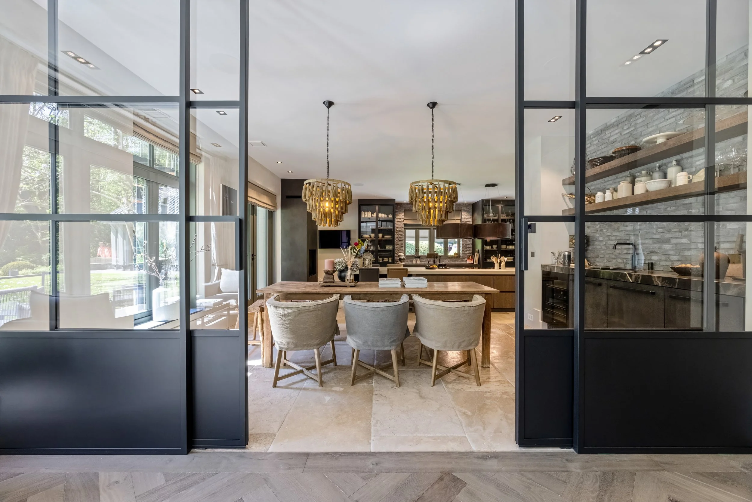 Modern keuken en eethoek met grote glazen schuifdeuren, houten tafel, beige stoelen en gouden hanglampen.