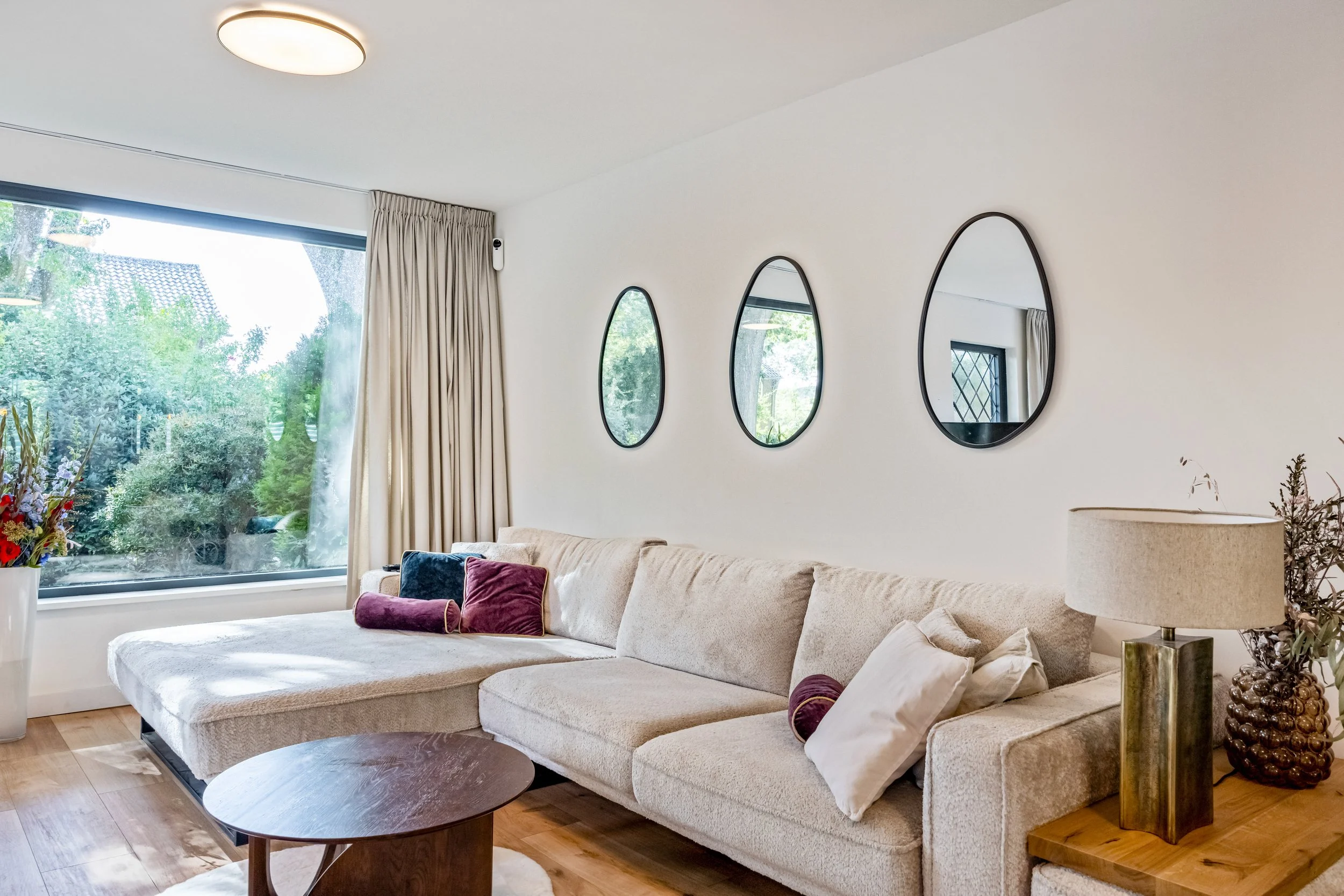 Woonkamer met grote raampartij, beige bank met kussens, kleine ronde houten salontafel, muur met drie decoratieve spiegels, lamp en bloemen op bijzettafel.