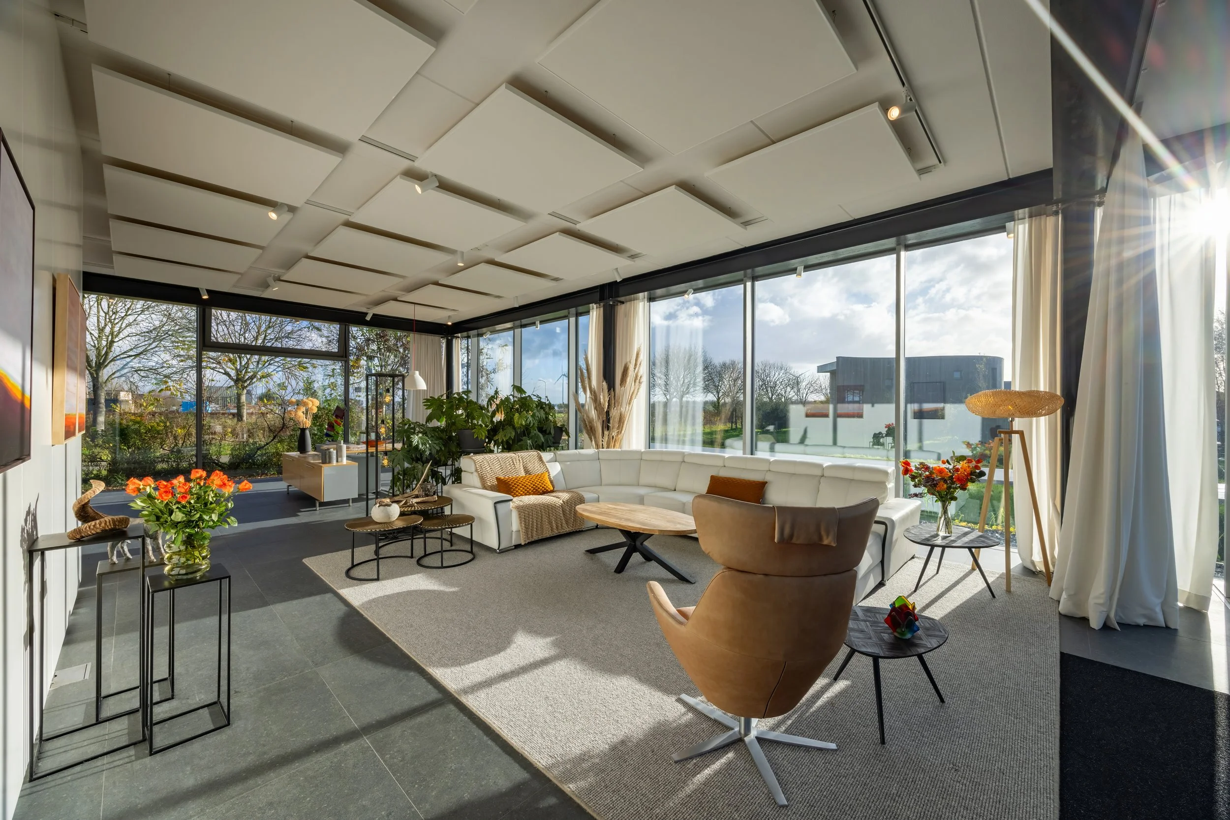 Lichte woonkamer met grote ramen, wit bank, fauteuil, salontafels, planten en decoraties, met uitzicht op de tuin en de straat.