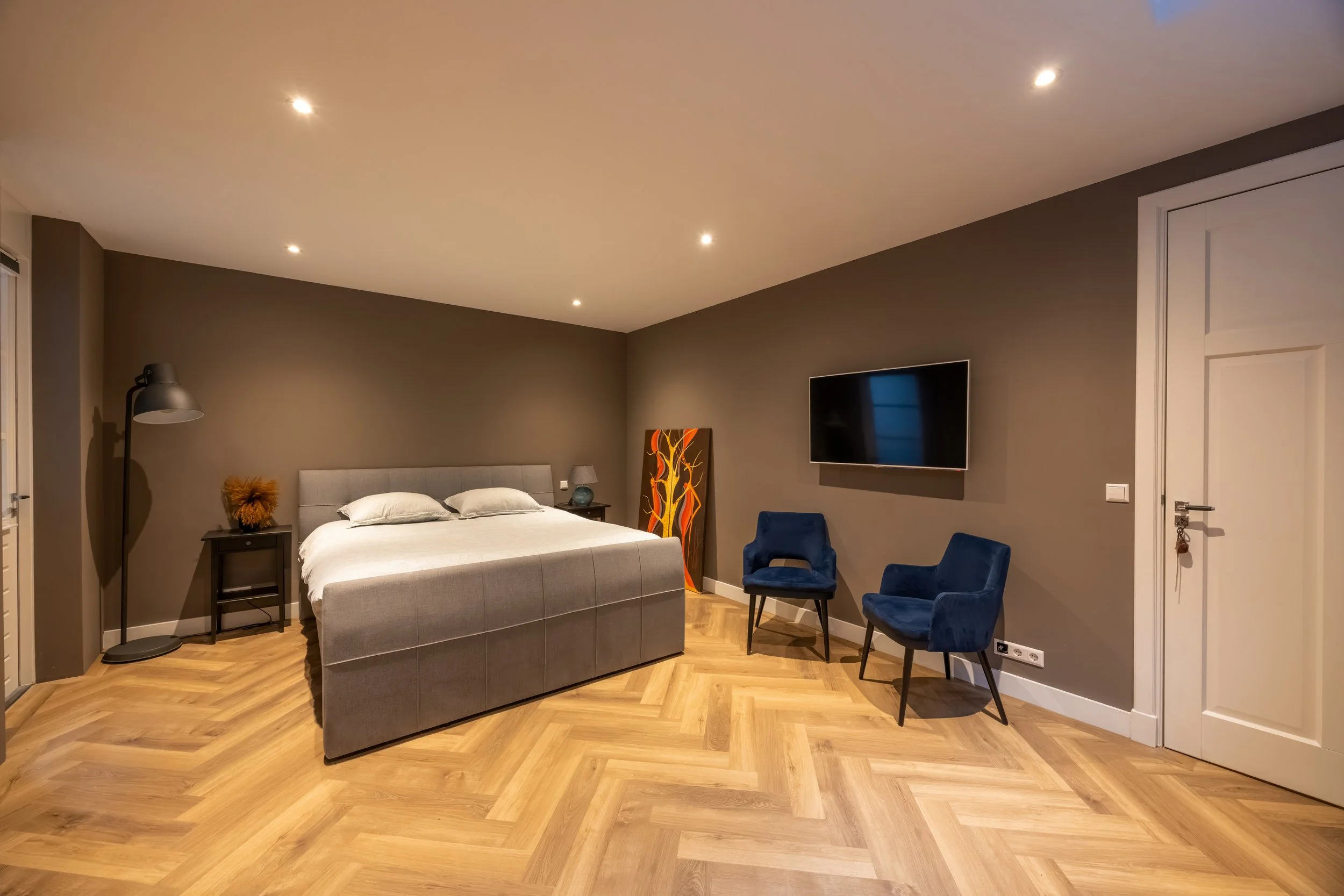 Modern hotelkamer met tweepersoonsbed, twee blauwe stoelen, wandtelevisie, houtenvloer, en decoratie.