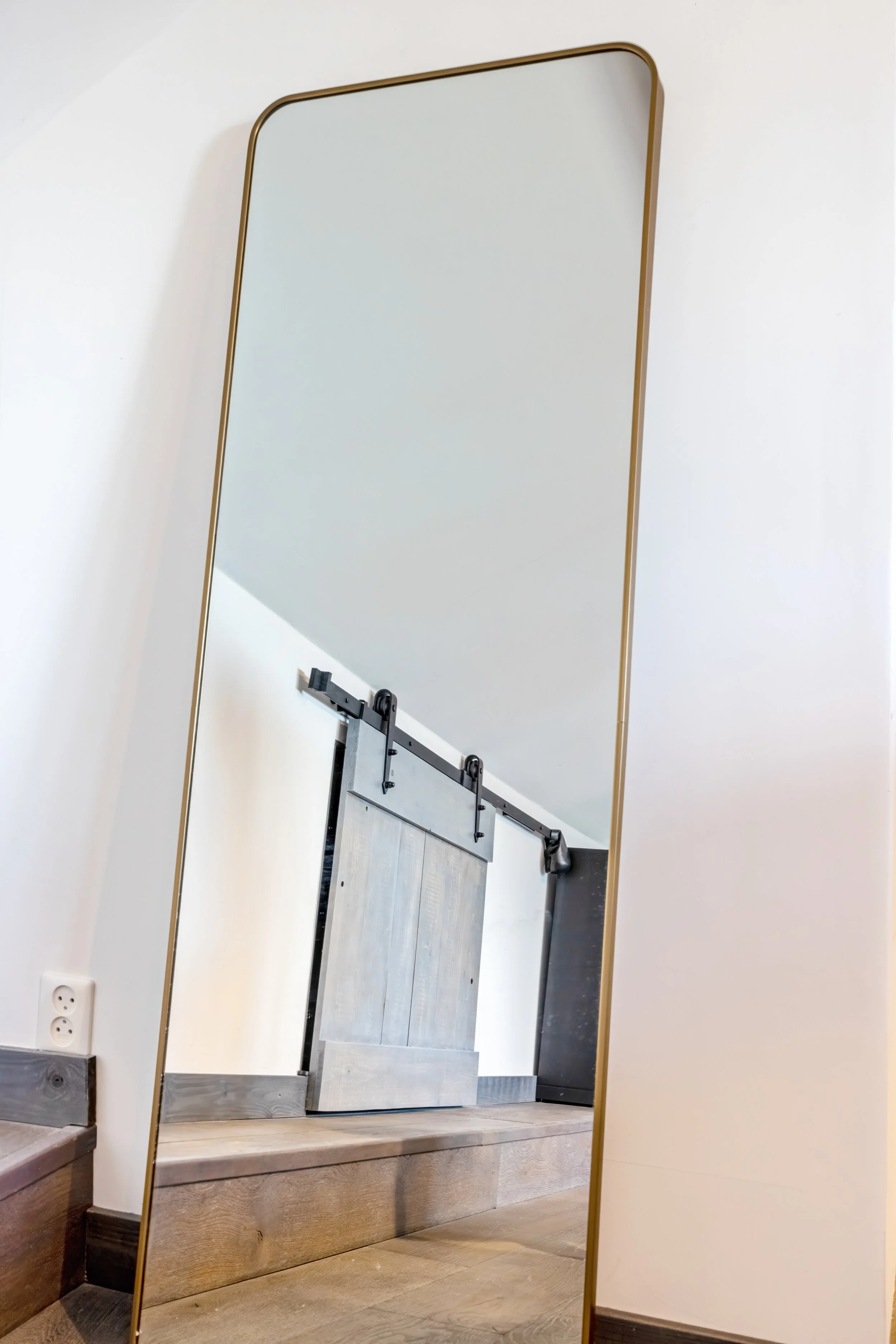 Een grote staande spiegel met een gouden rand, die een trap en een schuifdeur met een roedel rail reflecteert.