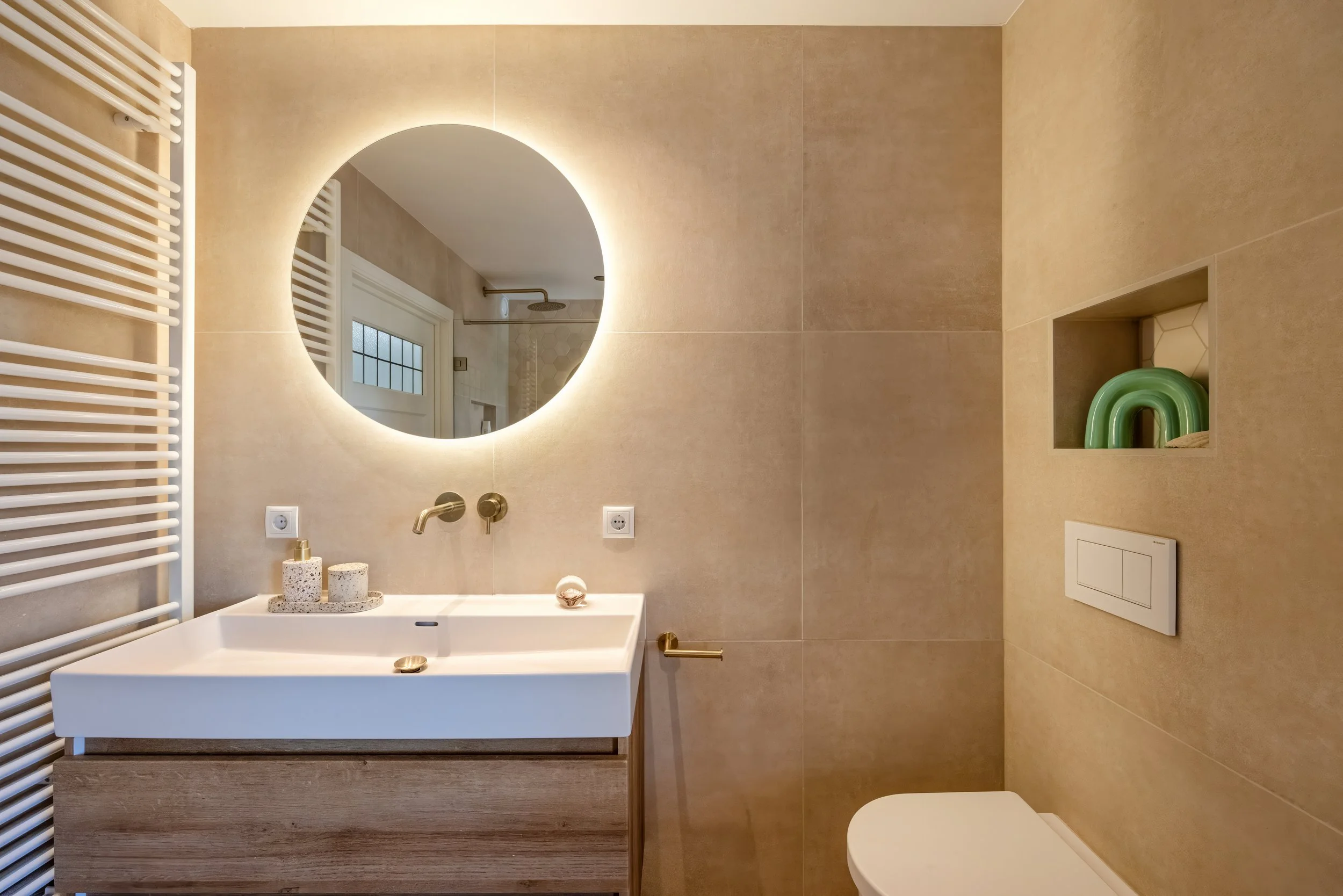 Moderne badkamer met ronde spiegel met verlichting, witte wastafel, muurmontuurkranen, een handdoekradiator, een ingebouwde nis met groene decoraties, en een wandtoilet.