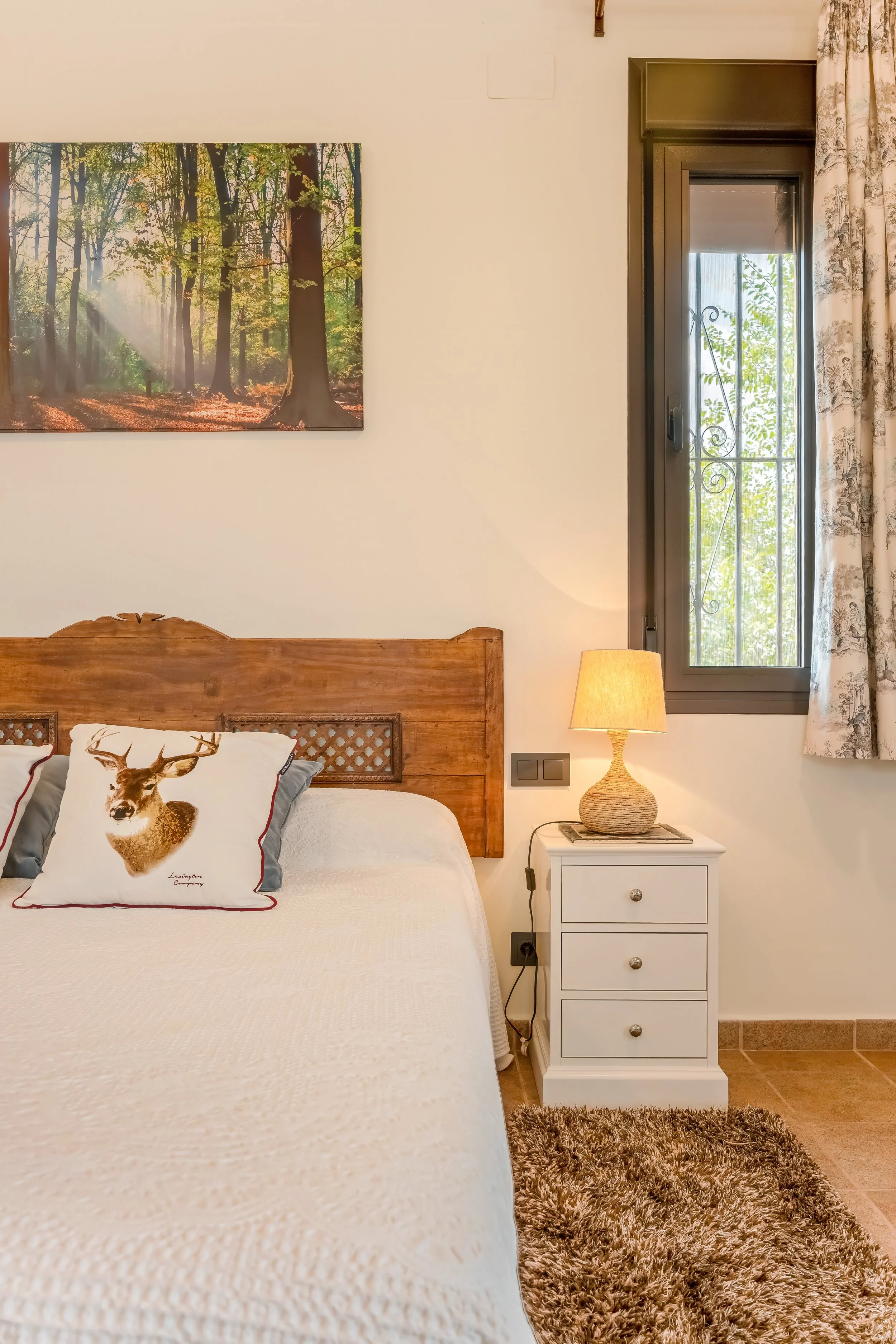 Kamer met wit bed, houten hoofdbord, sierkussen met hert, nachtkastje met lamp, raam met sierijzer, bosachtig schilderij