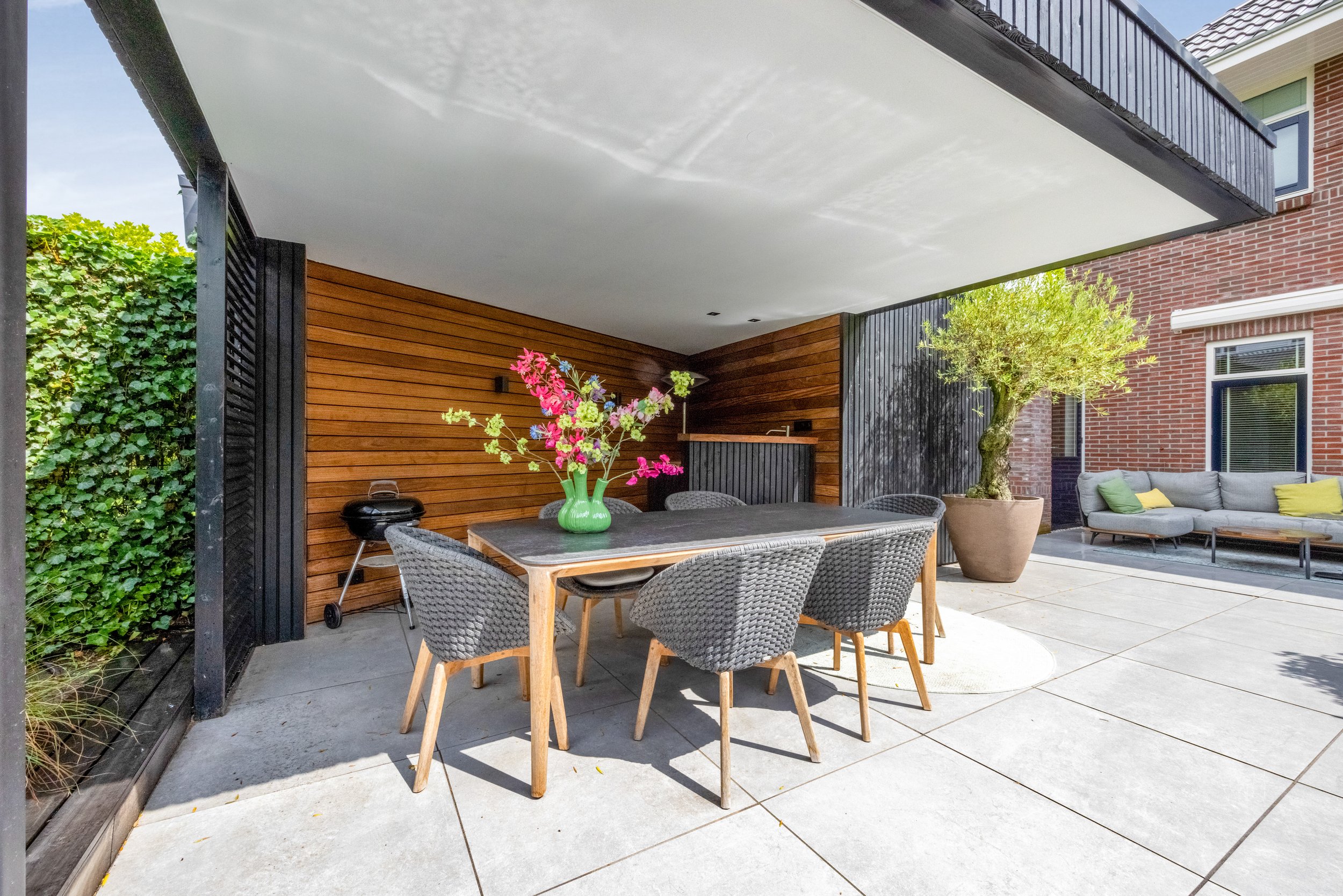 Moderne patio met houten en zwart decor en een eettafel met een bloemenvaas in het midden, buitenbank met kussens, en een grote pot met een boom.