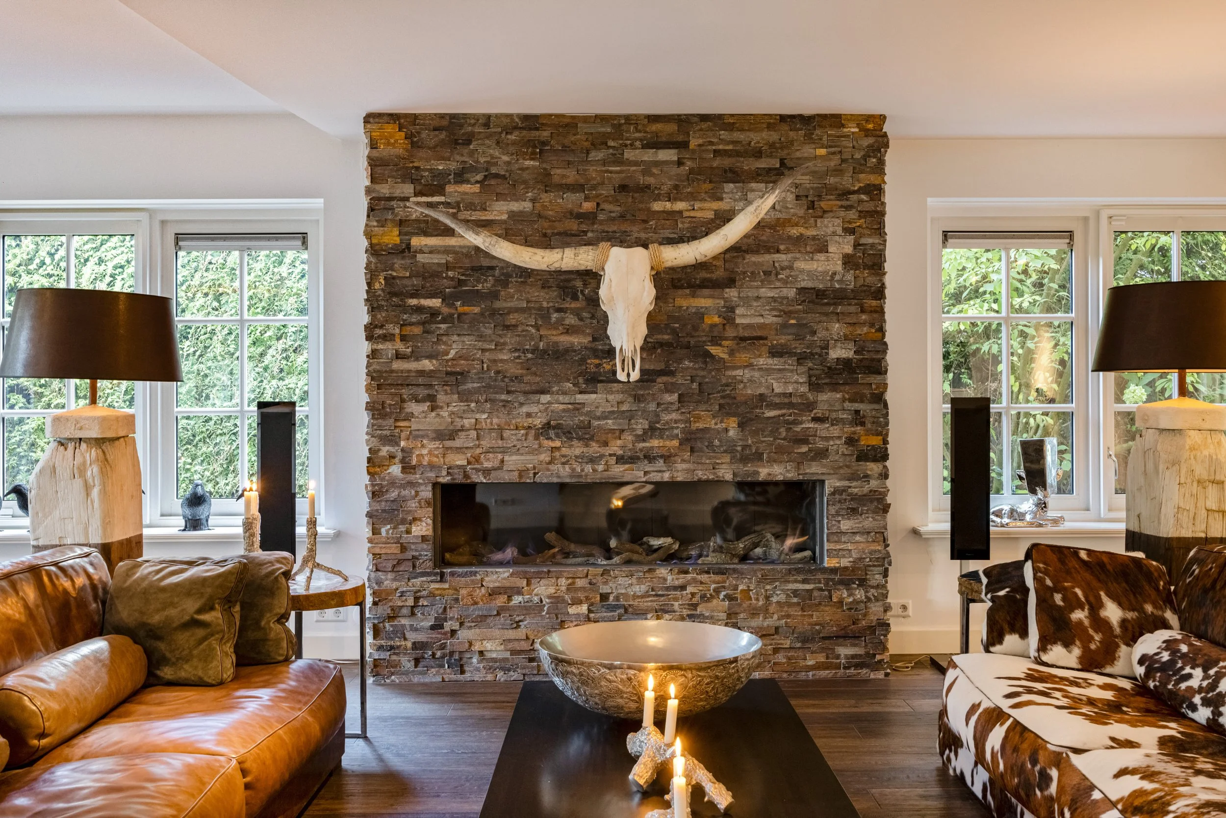 Interieur van een woonkamer met een bakstenen open haard, een schedel van een dier met lang gebochelde horens boven de haard, twee stoffen banken, een tafel met kaarsen en een grote urn in het midden.