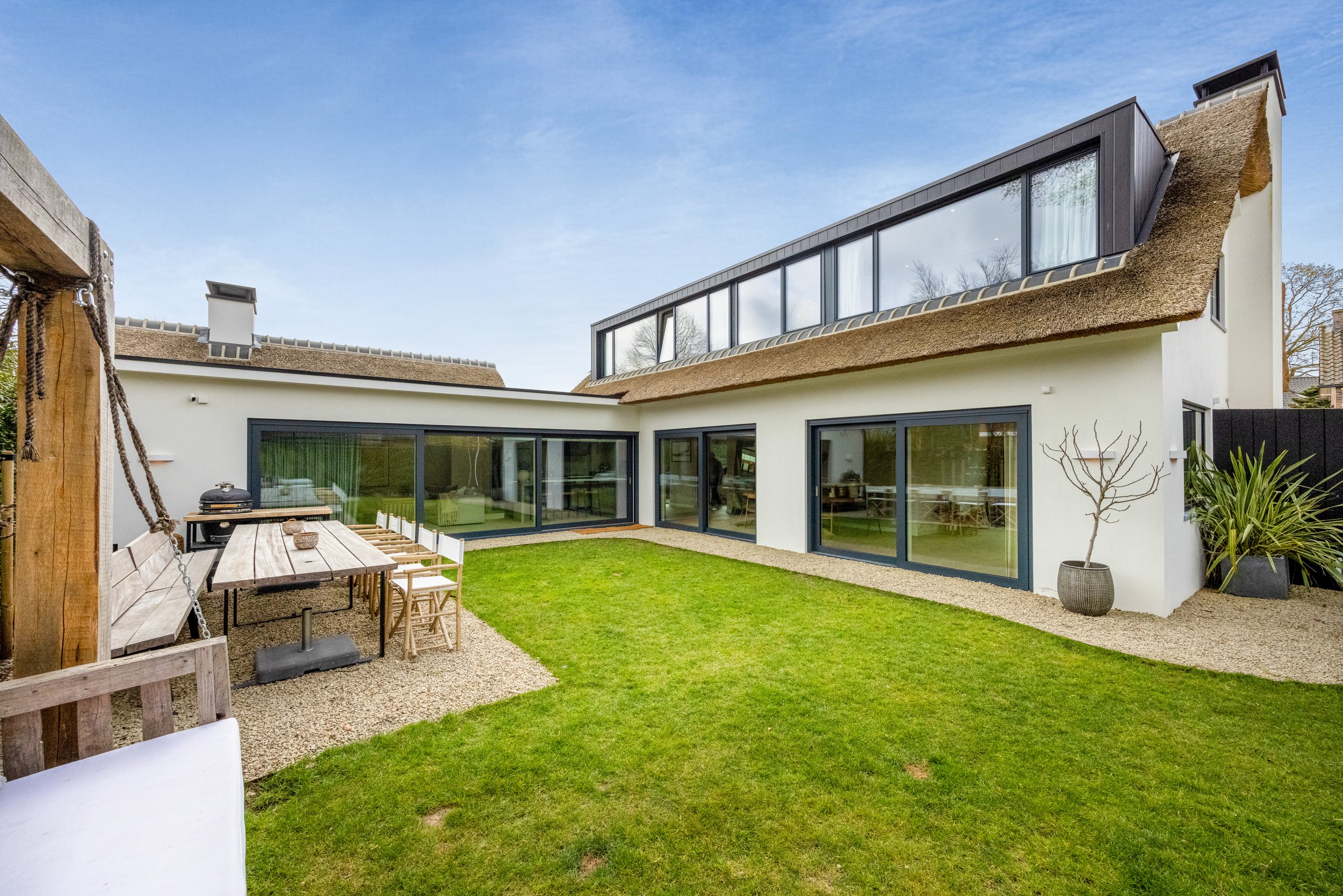Moderne witte woning met grote ramen, dat uitkijkt op een groene tuin met terras en zitplek, onder een blauw hemel.