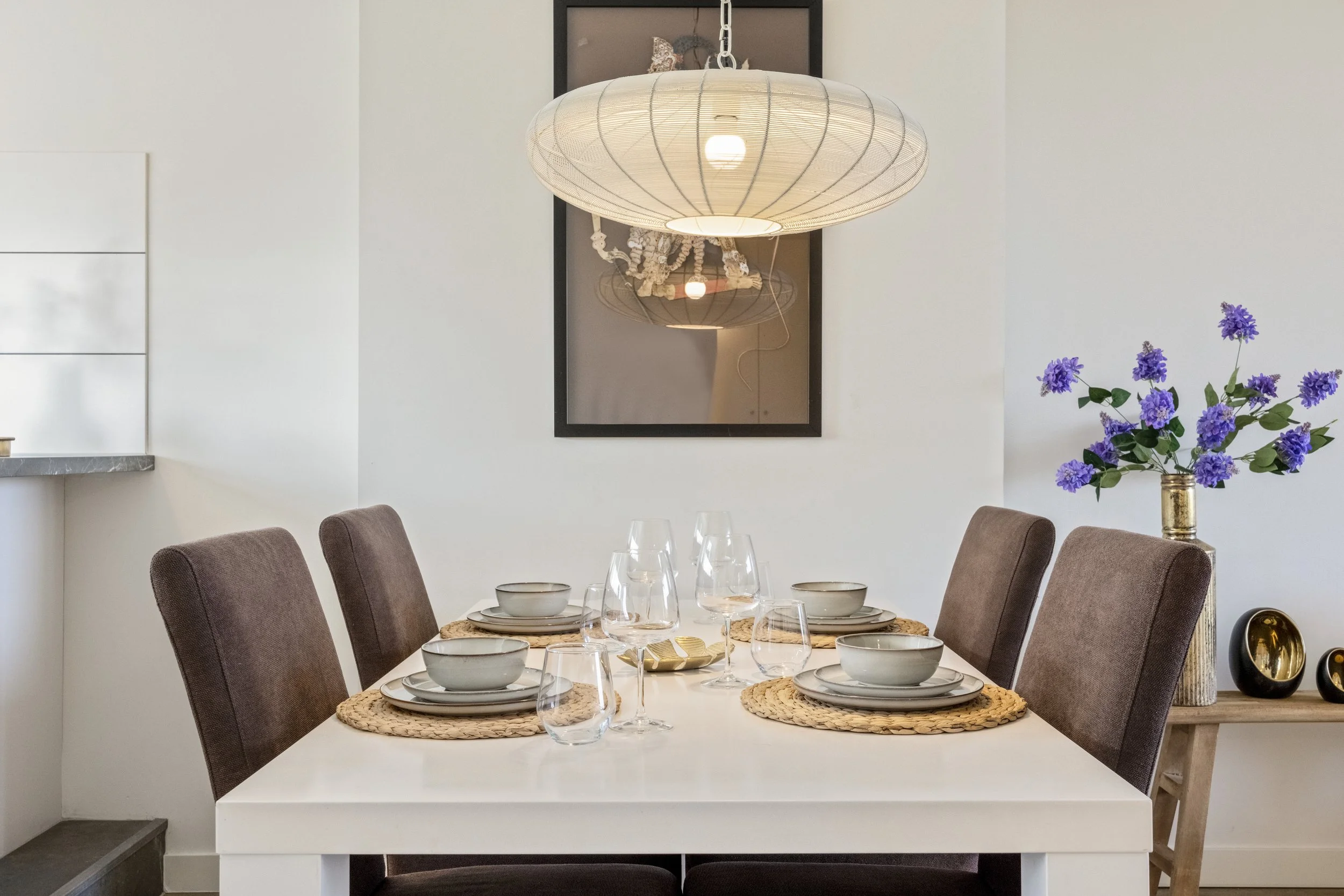 Eetkamertafel met zes borden, glazen en kommen, boven een lamp en een spiegel met een afbeelding van de lamp, in een kamer met witte muren en een kast met bloemen.