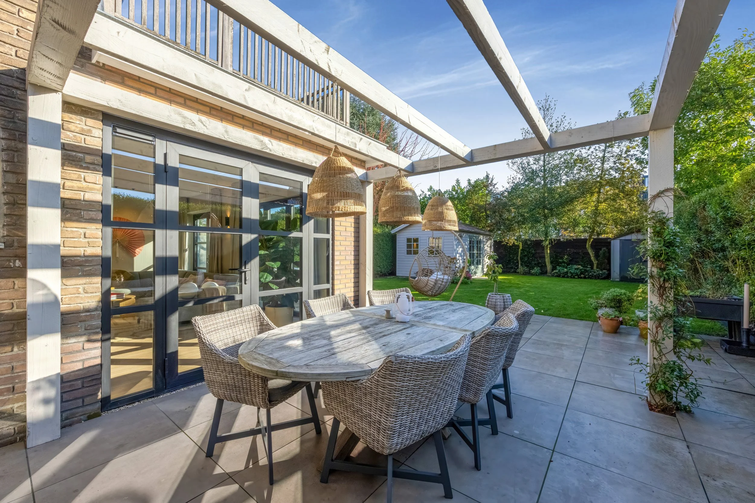 Een tuinterras met een houten tafel en acht rieten stoelen, onder een houten pergola met hangende rieten lampenkappen, uitkijkend op een groene tuin met een gazon en een schommelstoel.